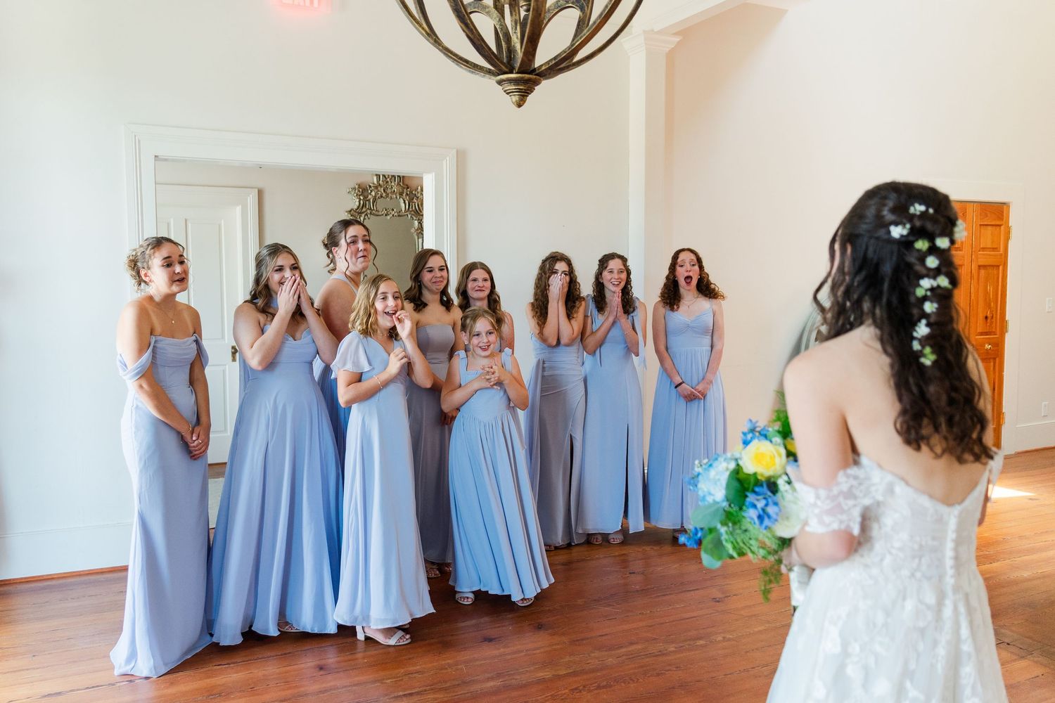Bride in lace gown holds bouquet during first look reveal with bridesmaids in blue dresses reacting with joy.
