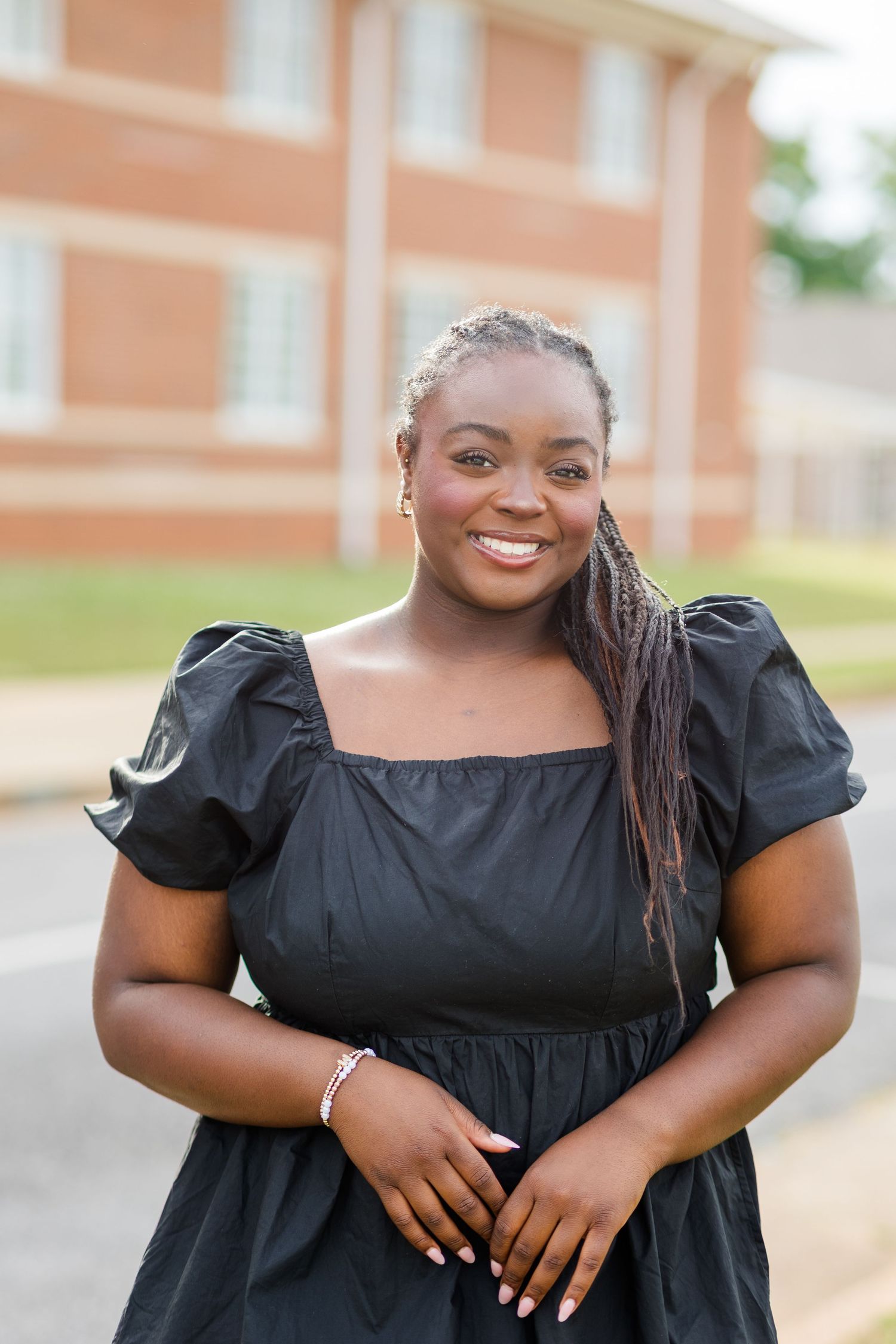 Smiling young Black woman in black puff-sleeve dress standing outdoors near brick building.