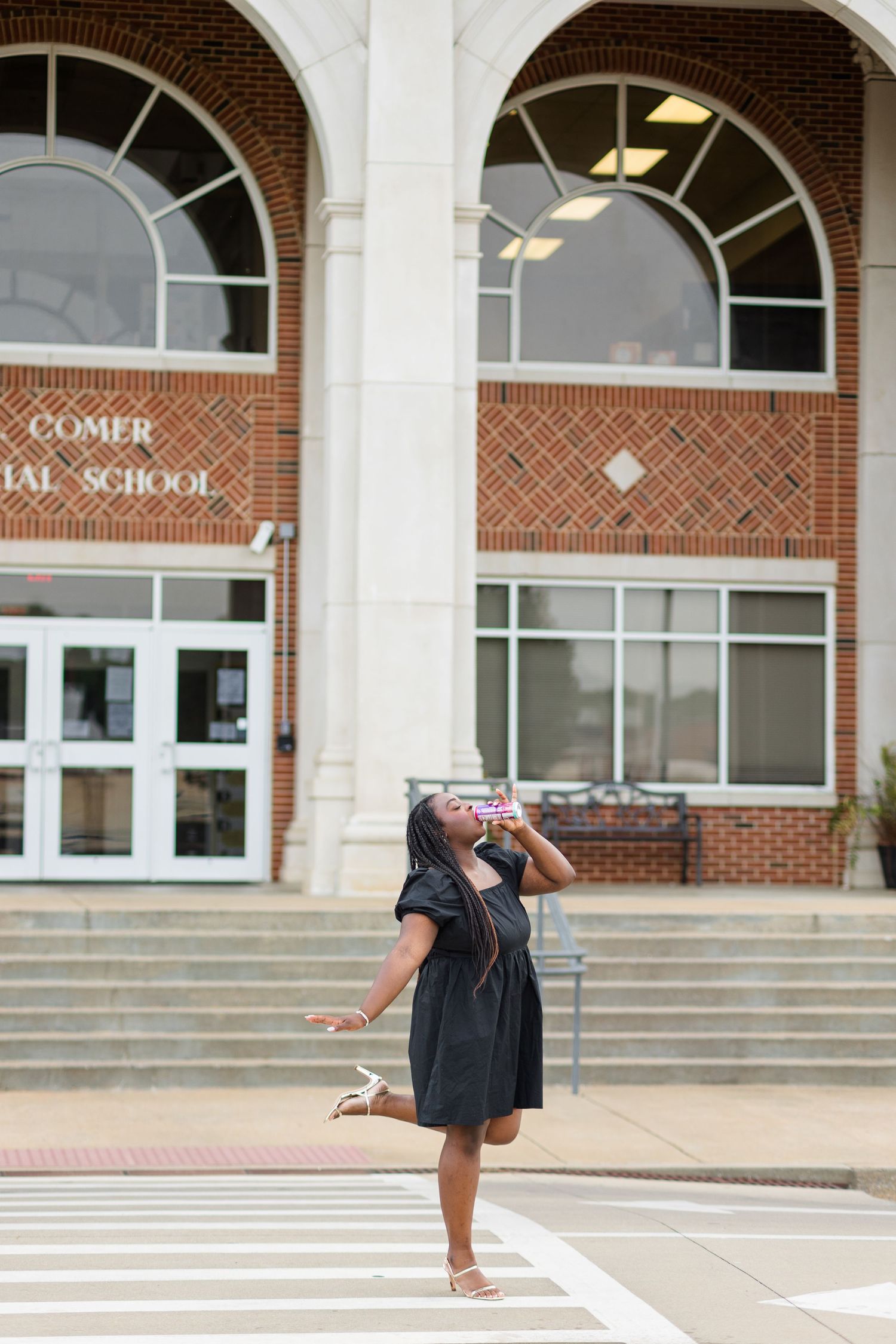 Joyful young woman in black dress celebrating in front of B.B. Comer Memorial School brick building.