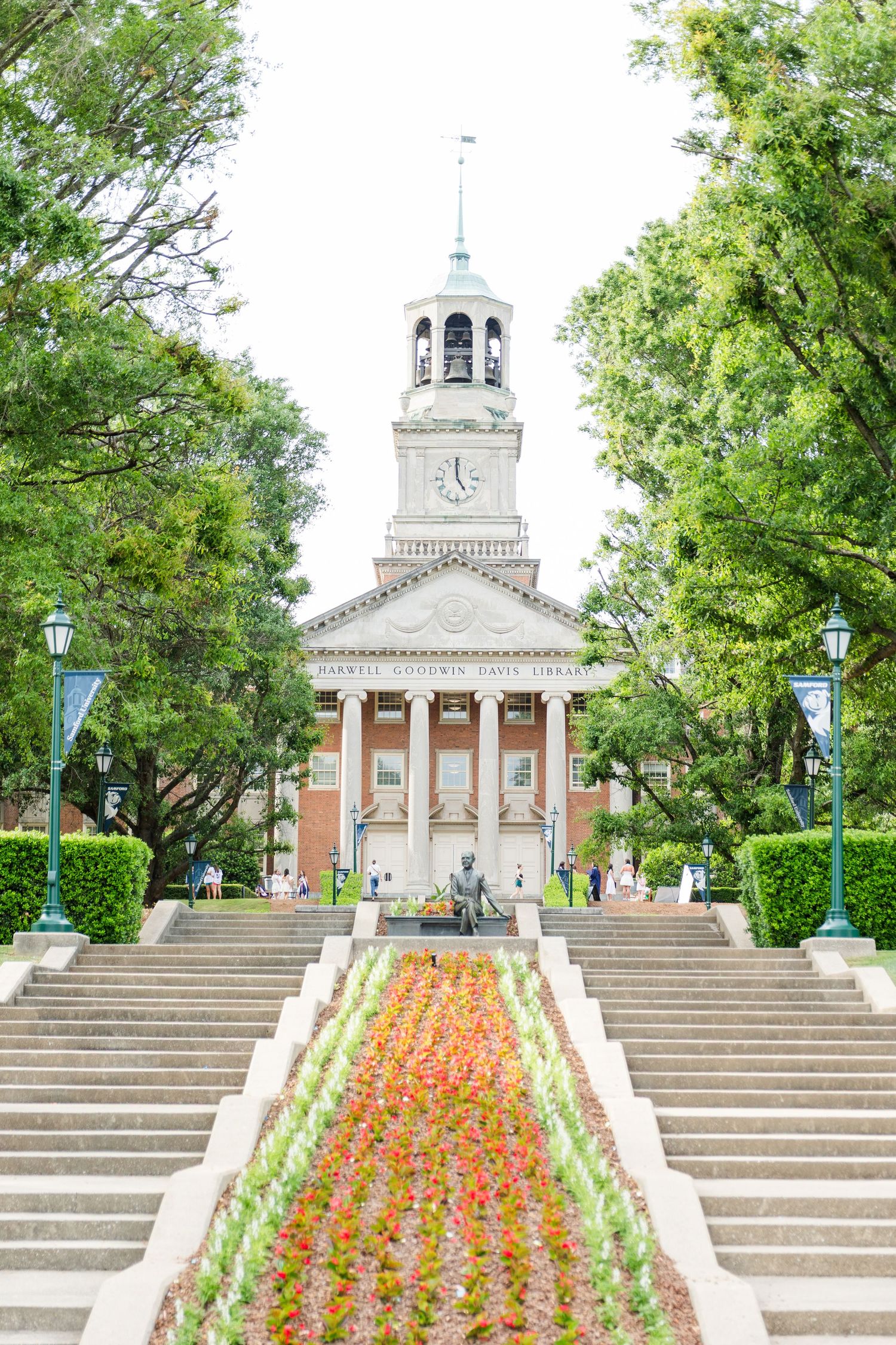 Historic university brick building with white bell tower, colorful flower beds, and grand staircase entrance.