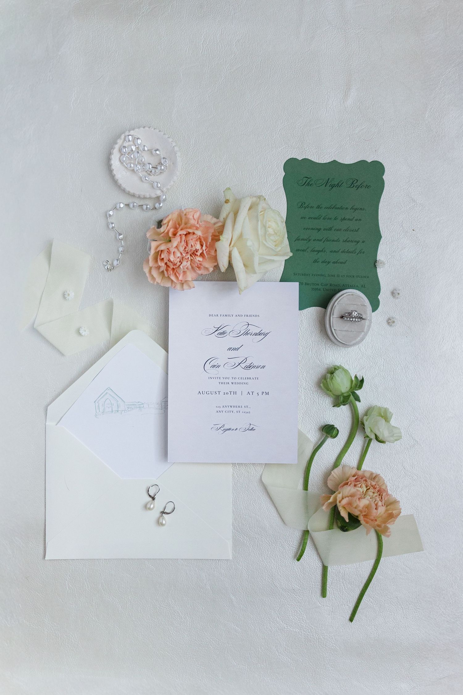 Elegant wedding invitation suite with green card, white envelope, pearl jewelry, and peach roses on a white background.