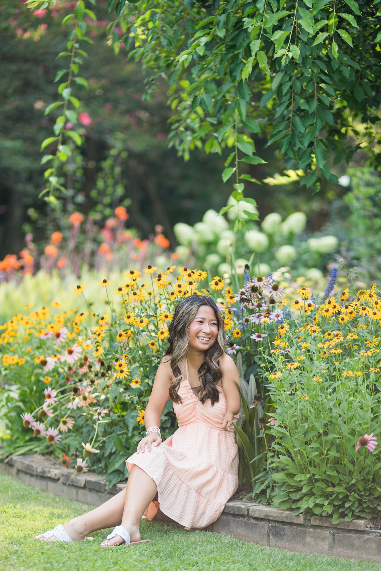Smiling woman in peach dress sitting in a vibrant garden with yellow, orange, and purple flowers.