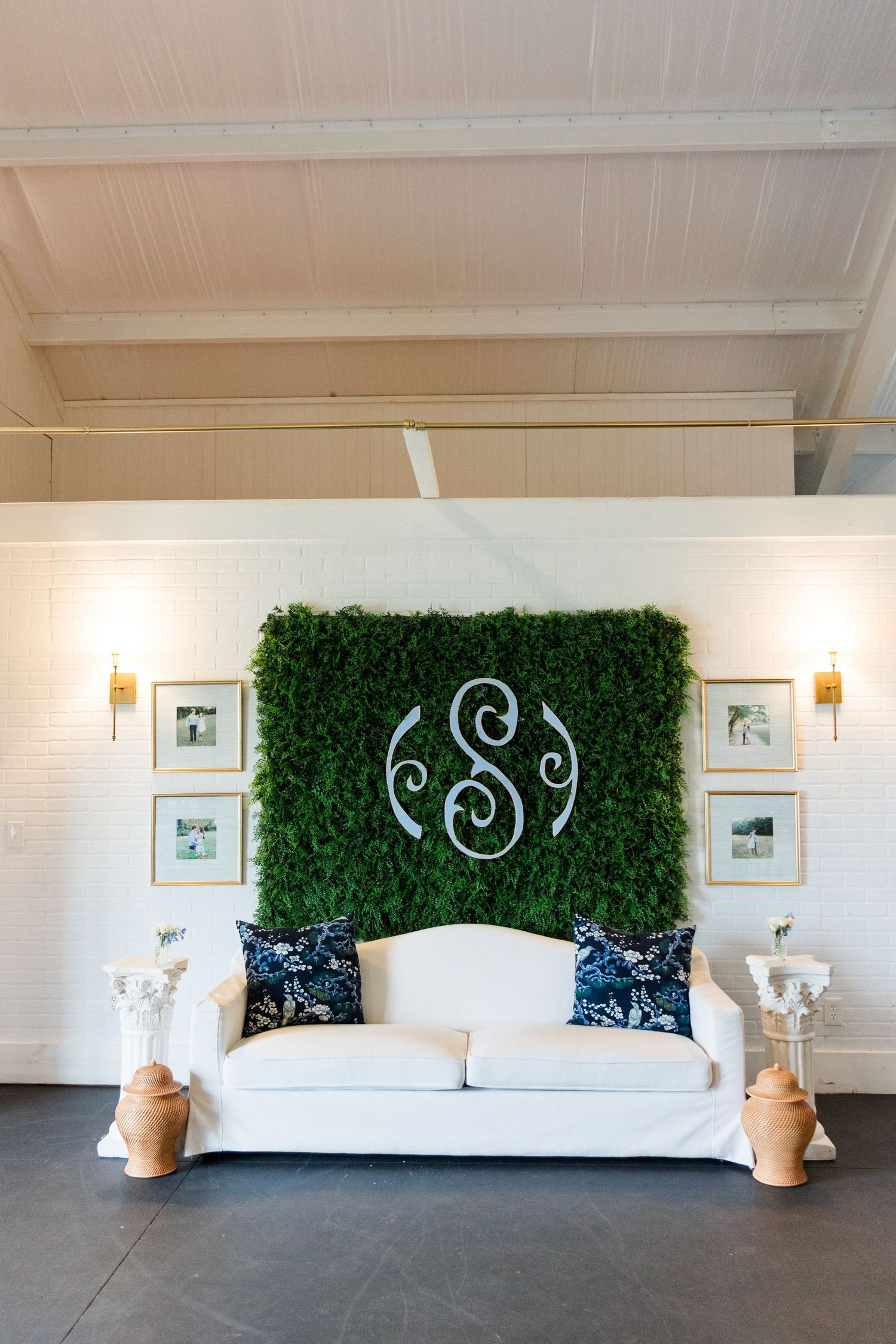 Elegant wedding lounge with white sofa, green boxwood monogram wall, and gold framed photos.