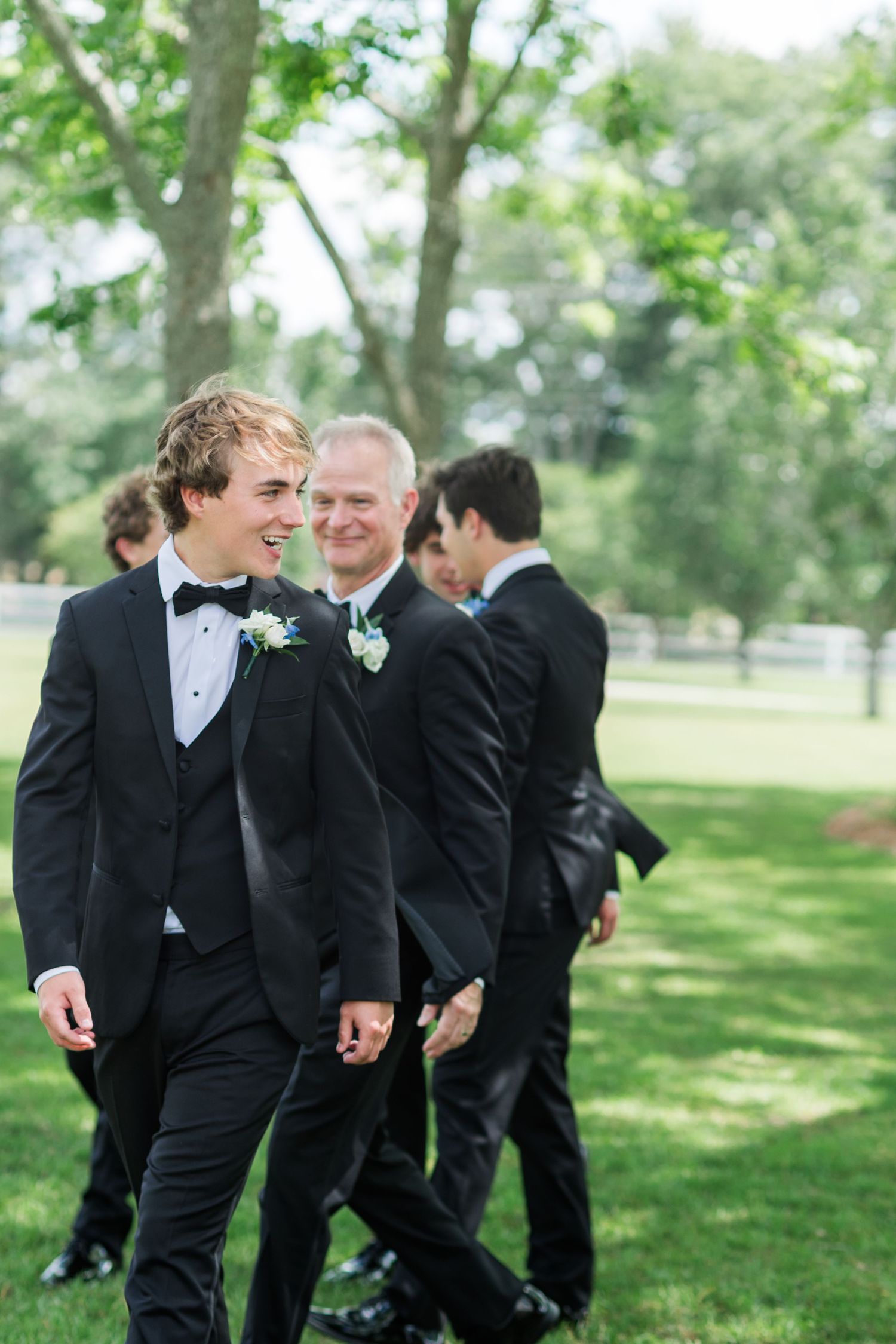 Groomsmen in black tuxedos walk together outdoors on green lawn during wedding celebration.