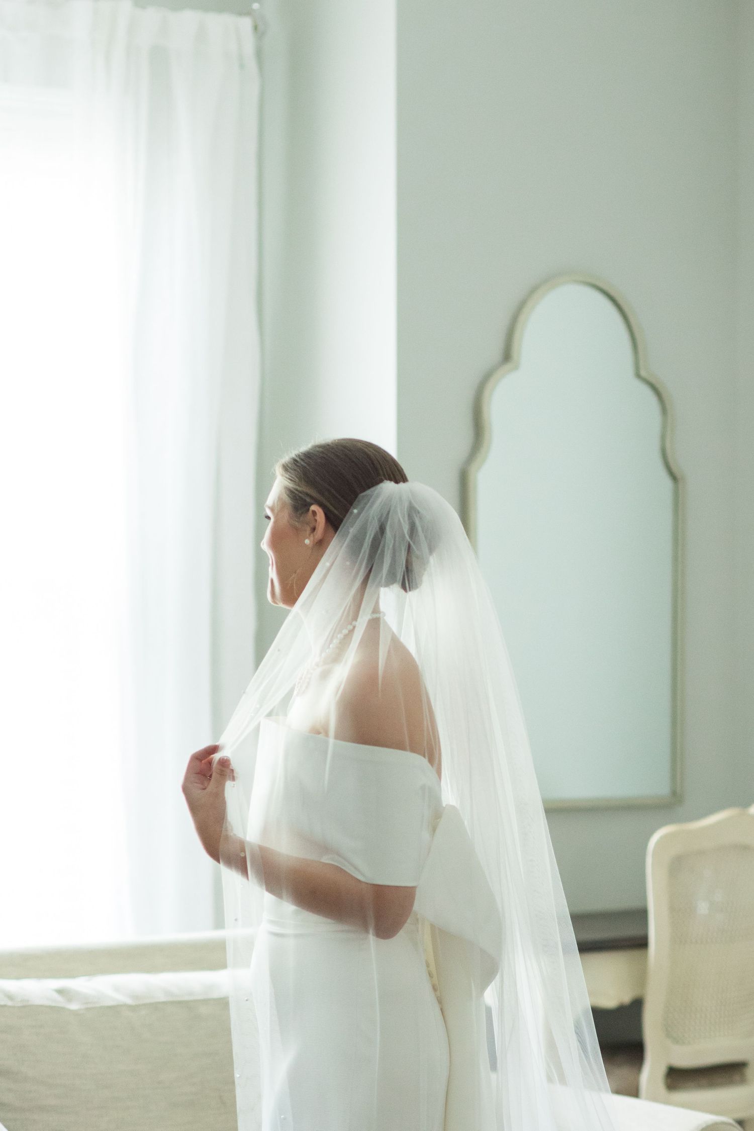 Elegant bride in off-shoulder gown and flowing veil standing by bright window in bridal suite.