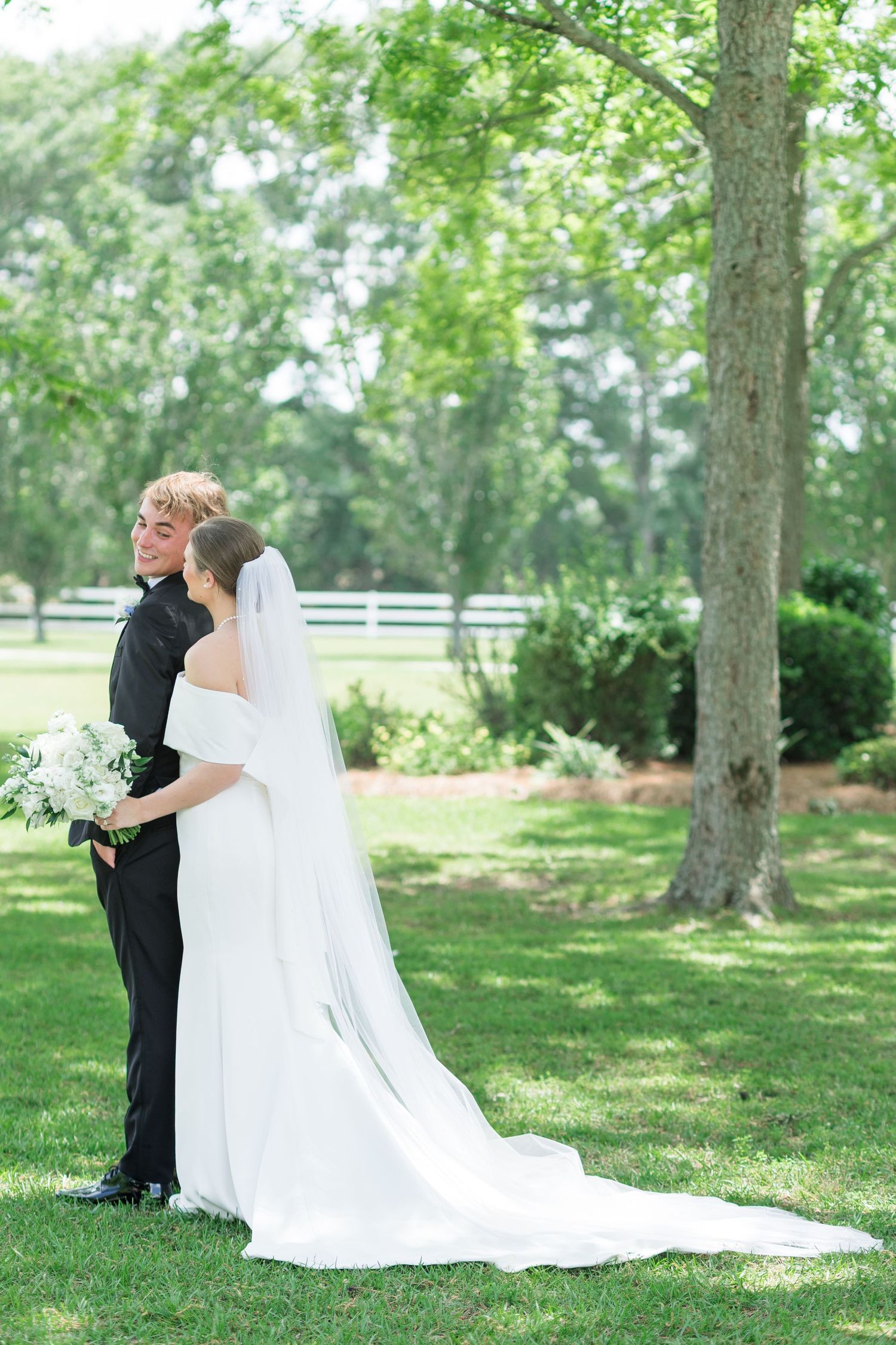 Bride and groom embrace outdoors on lush green lawn, bride wearing elegant white gown with long veil and bouquet.