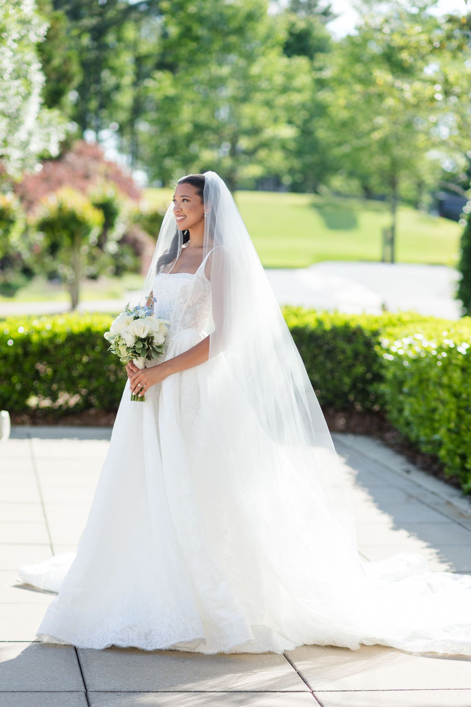 Bride in white ball gown with veil holding bouquet outdoors on sunny day.