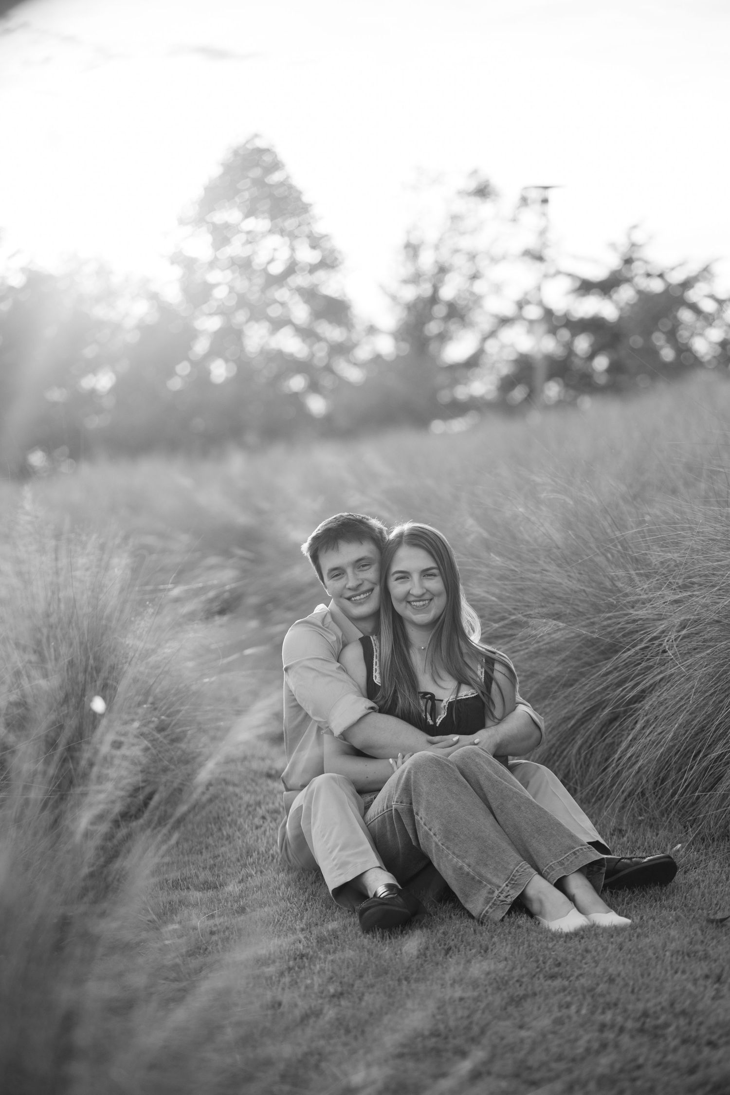 A couple sits together in black and white, embracing in a field of tall grasses with soft backlight.