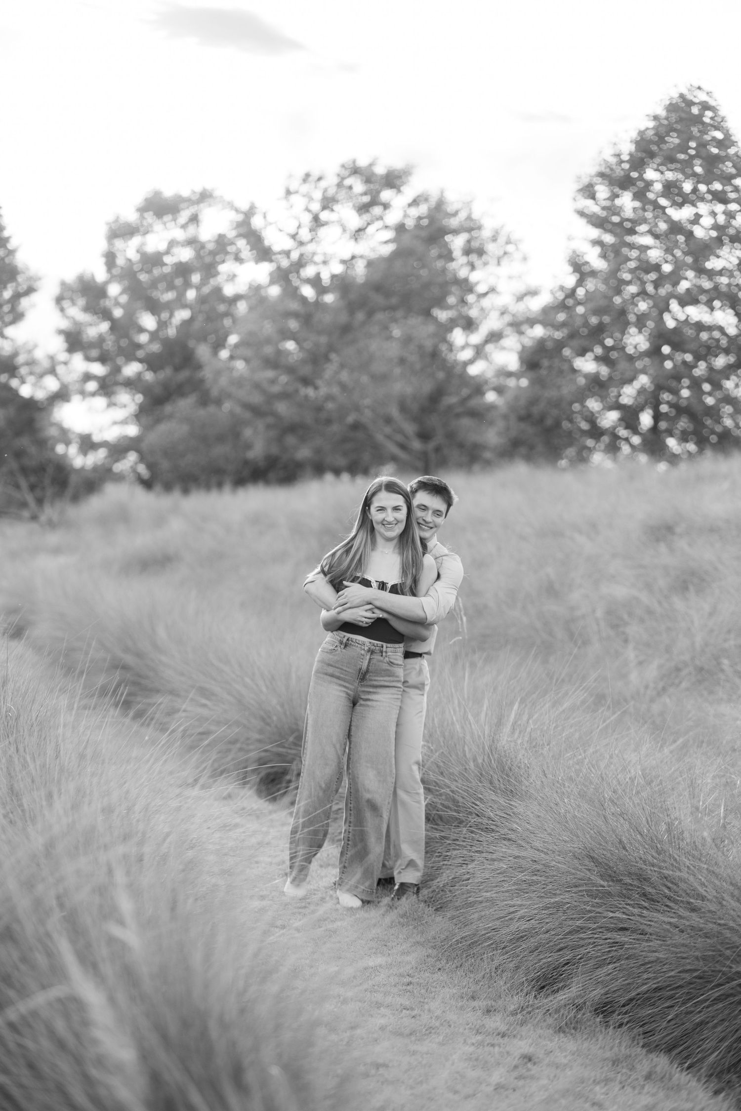 Black and white photo of a couple embracing on a grassy path surrounded by tall grass and trees.