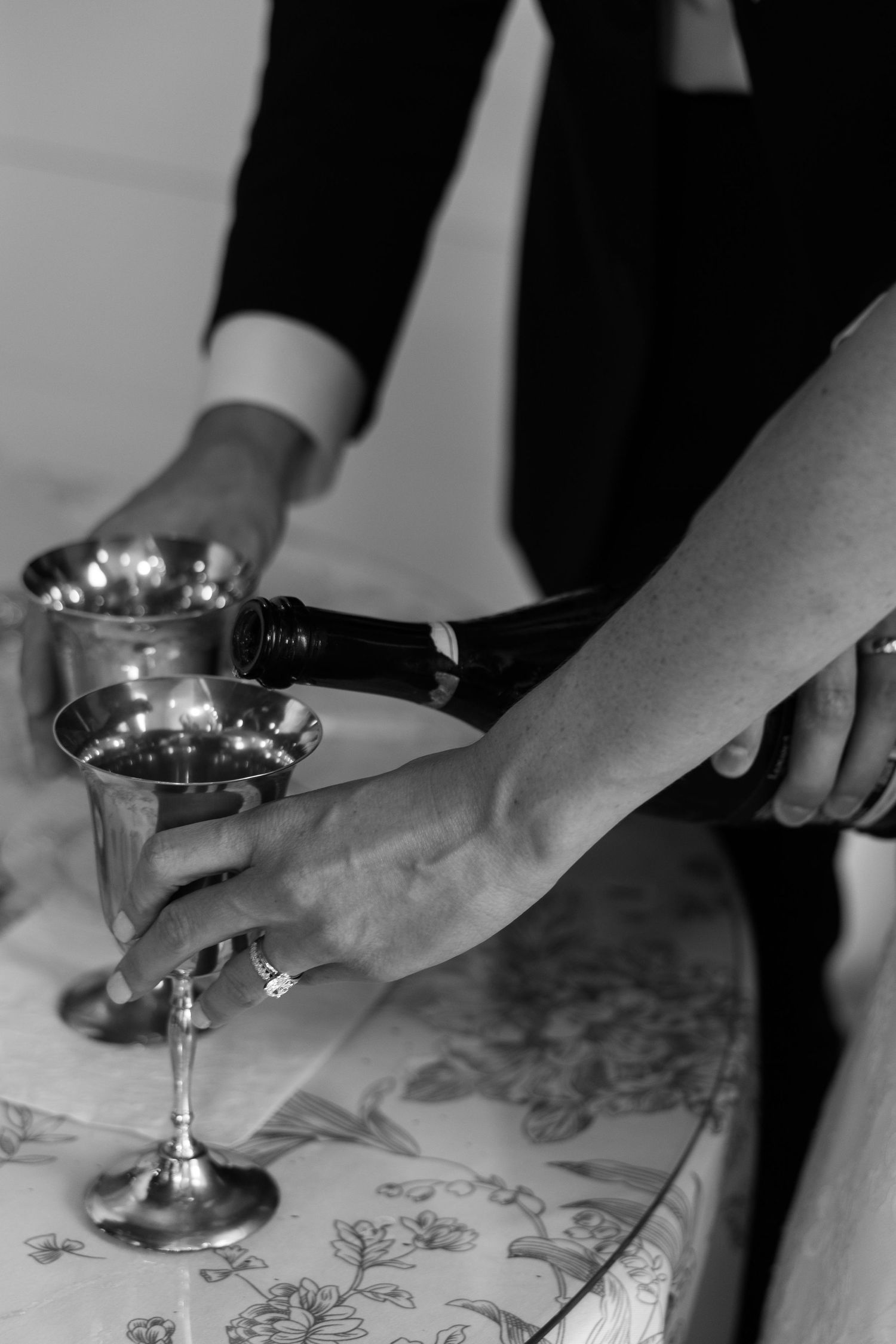 Black and white photo of hands pouring wine into silver goblets at a wedding ceremony.