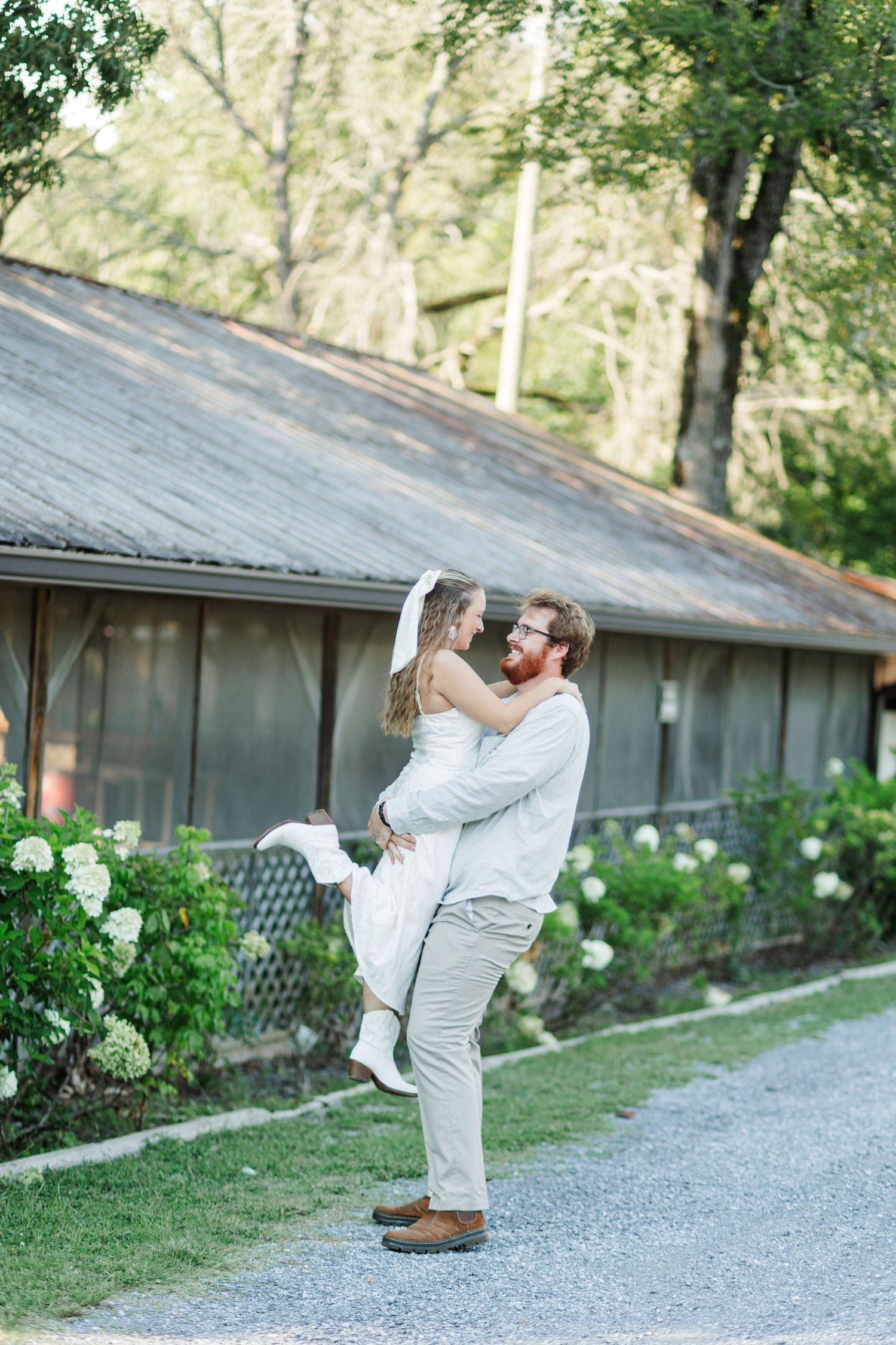 Groom lifts bride in white dress outdoors near rustic barn surrounded by lush greenery.