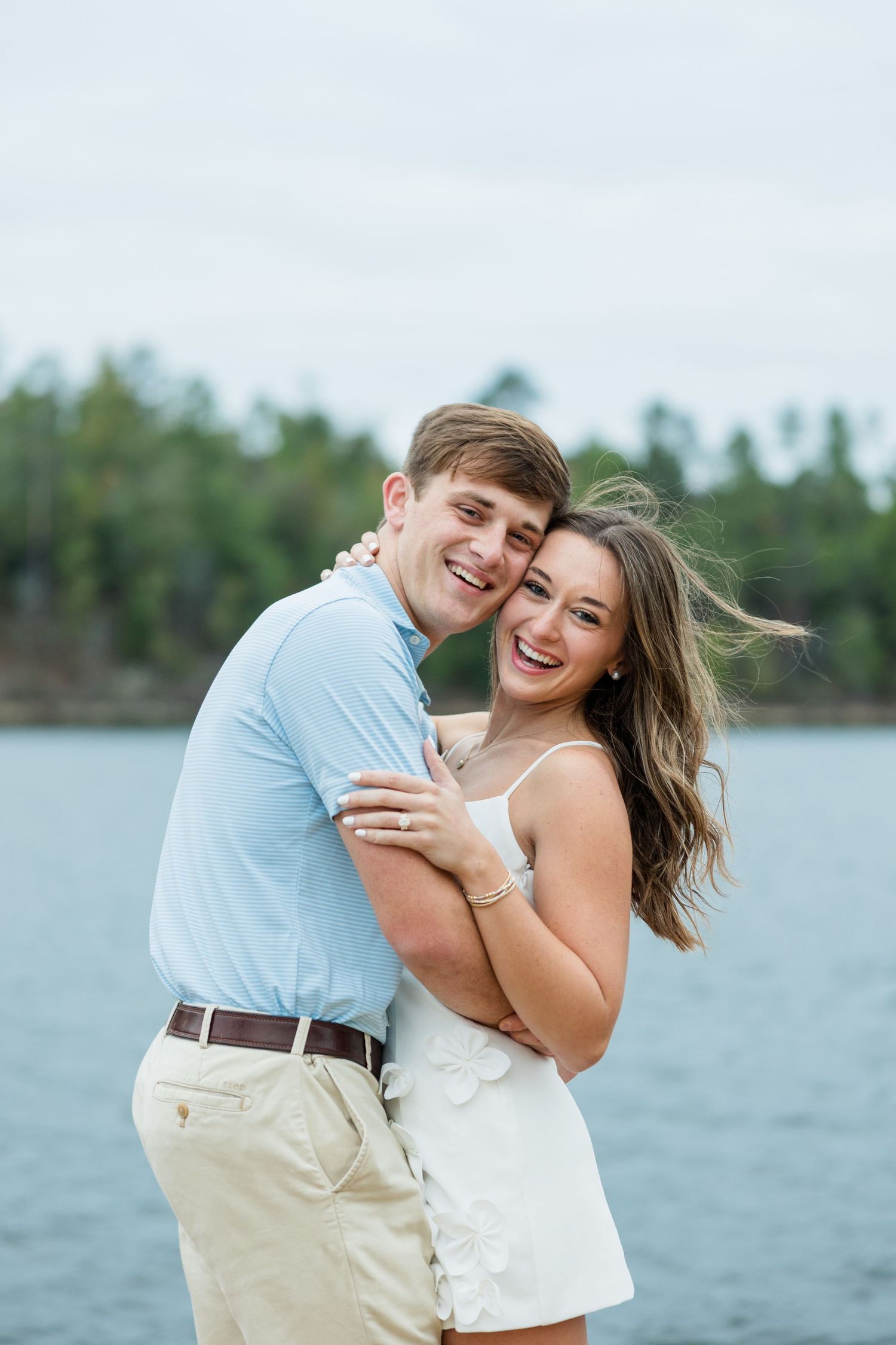 Smiling couple embracing in a joyful engagement photo beside a calm lake with a tree-lined shoreline under an overcast sky.