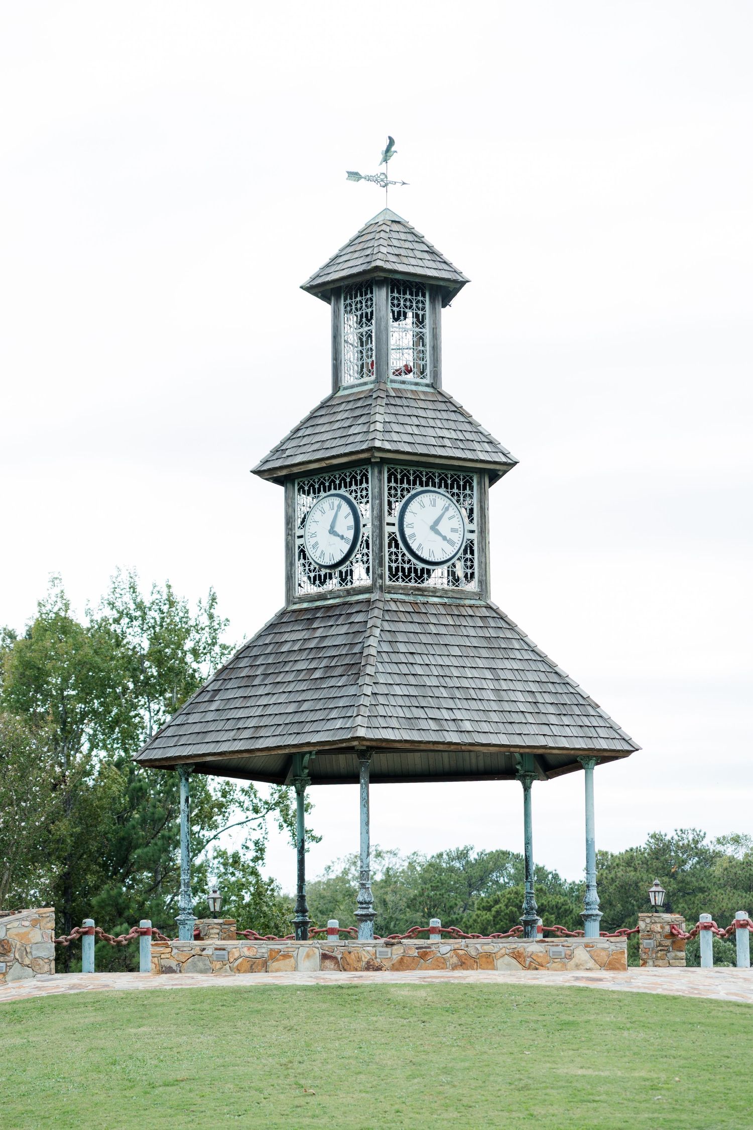 Tiered wooden clock tower with shingled roof, two clock faces and weather vane, standing on stone pillars beside a green lawn.