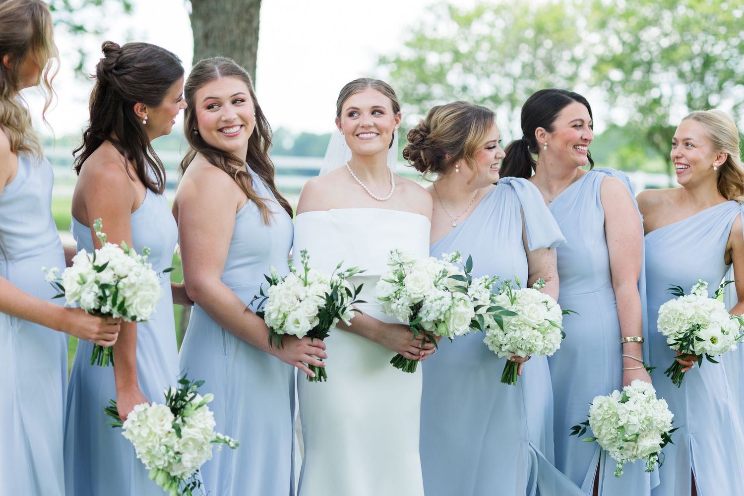 Bride in white gown with bridesmaids in light blue dresses holding white floral bouquets at outdoor wedding.
