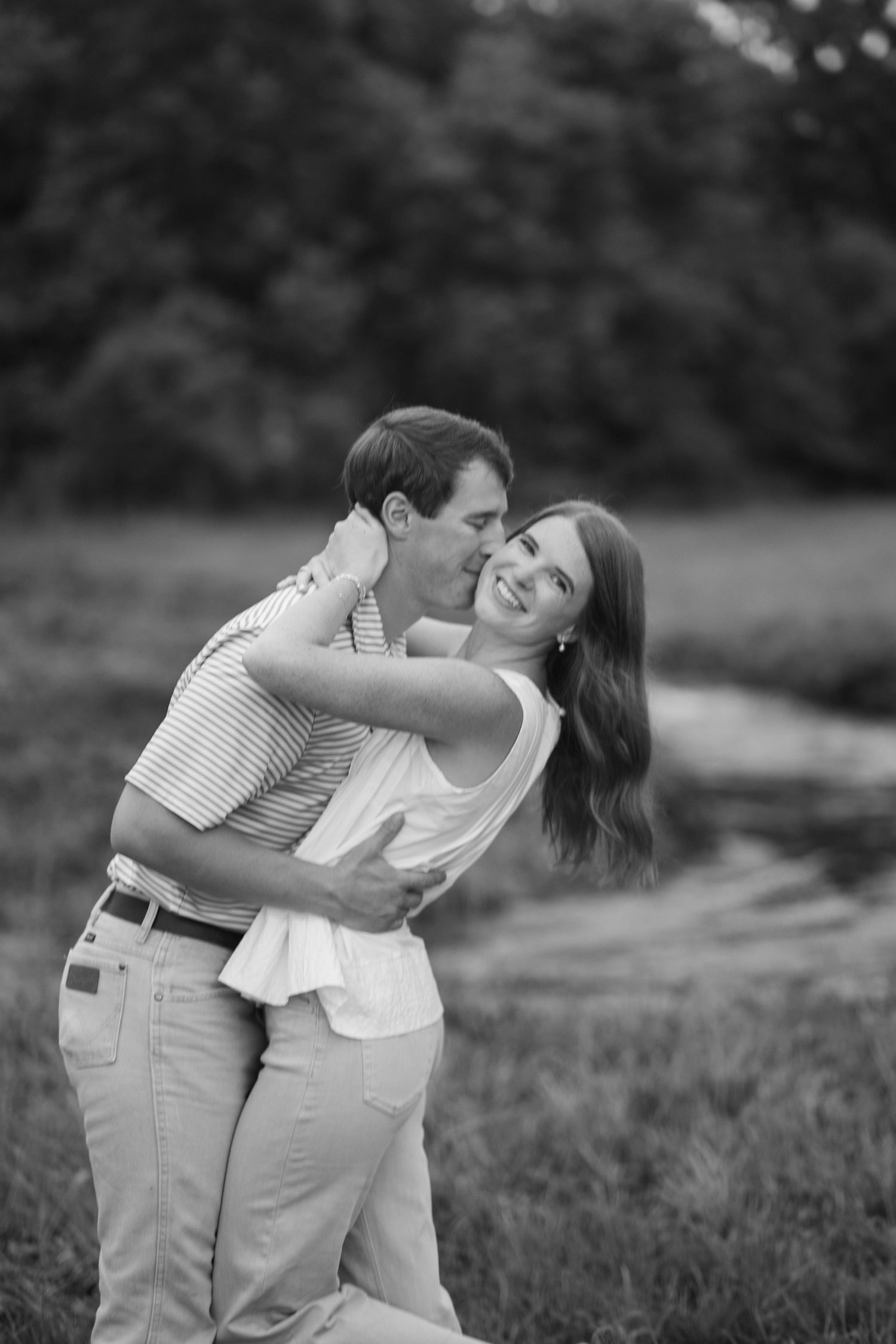 Black and white photo of a man kissing a laughing woman while embracing in a field.
