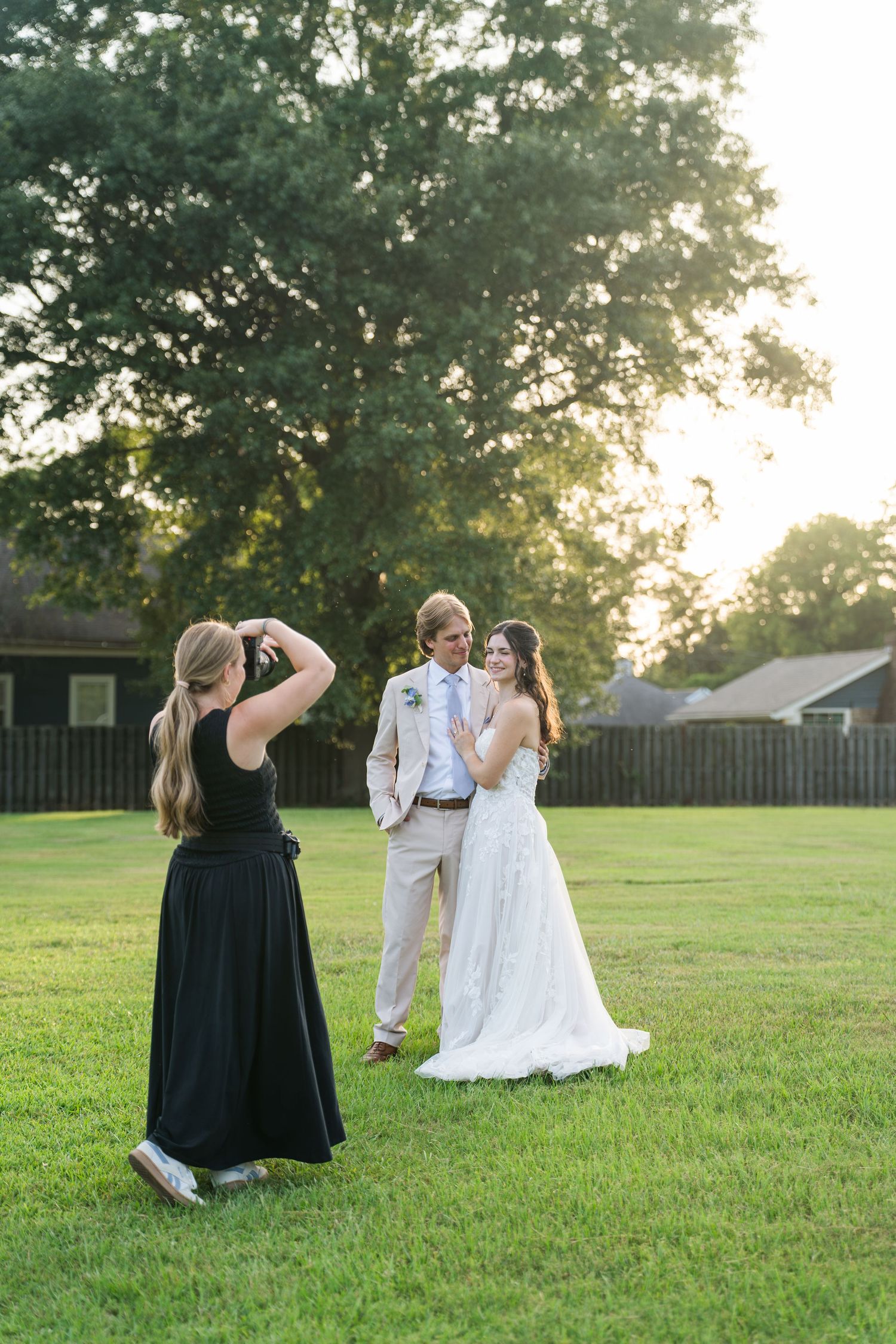 Photographer capturing bride and groom portraits outdoors on green lawn at golden hour sunset near wooden fence.