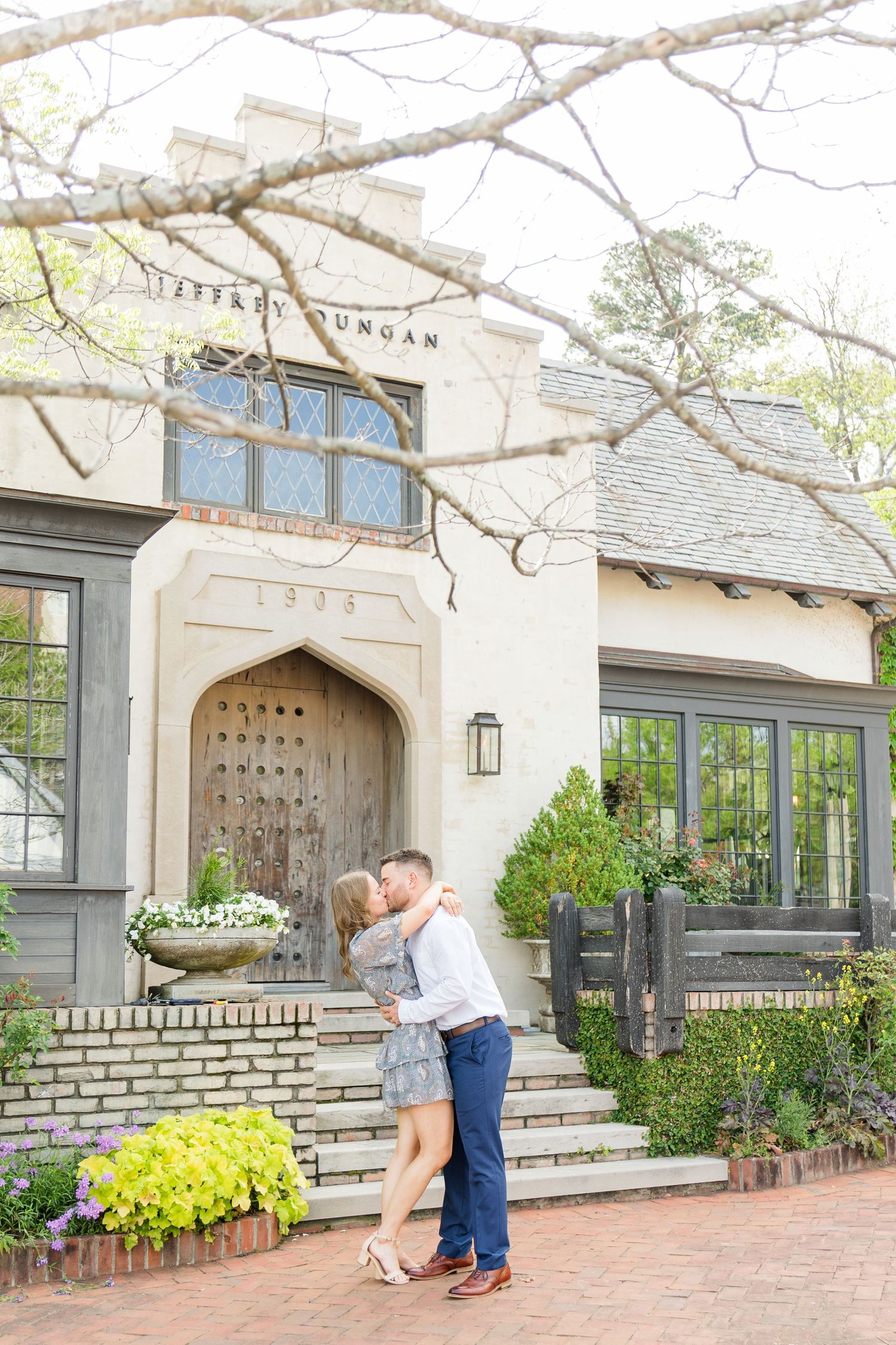 Couple sharing a romantic kiss in front of a charming Tudor-style building entrance surrounded by lush greenery.