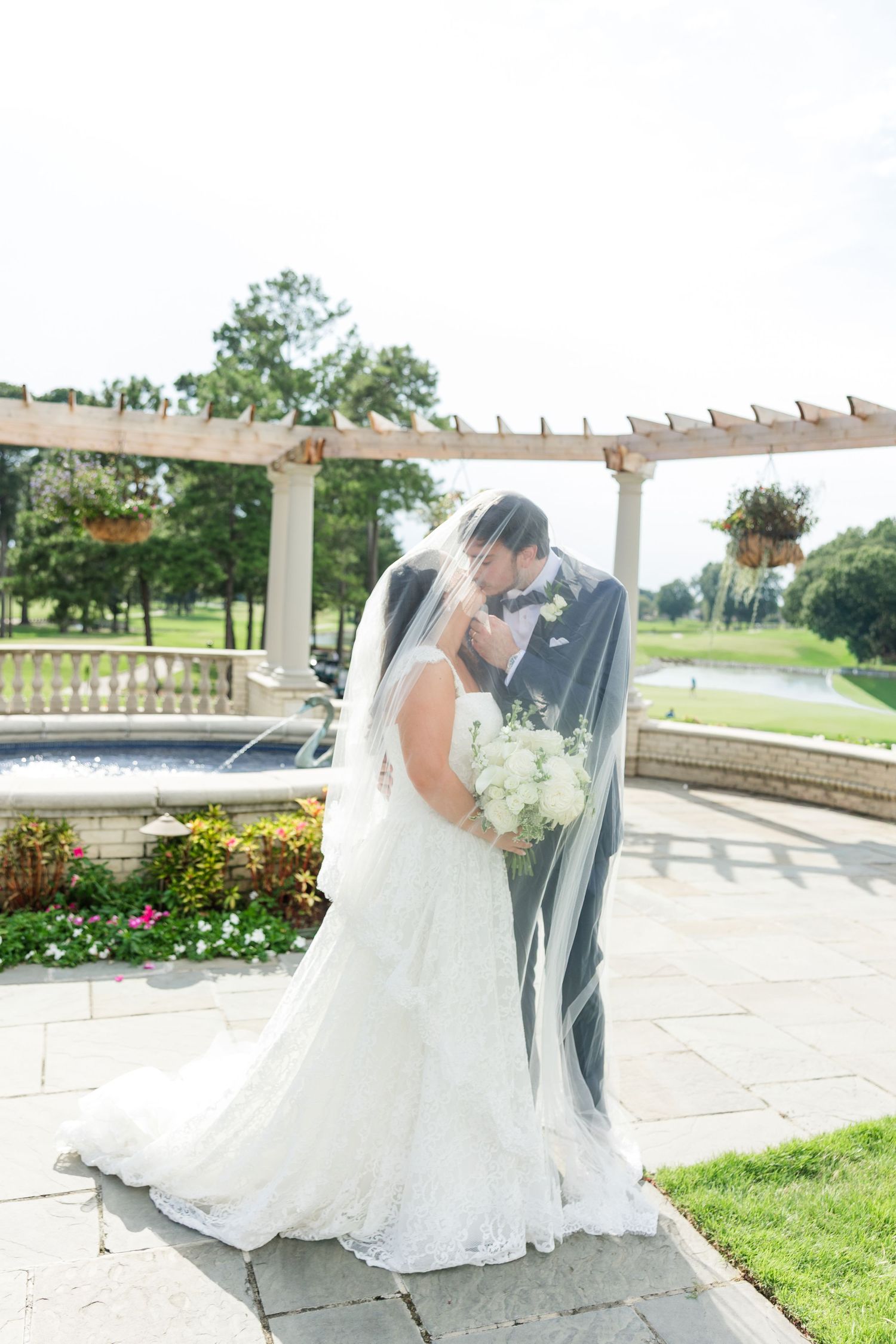 Bride and groom kissing under flowing veil beside fountain in elegant outdoor garden wedding setting.