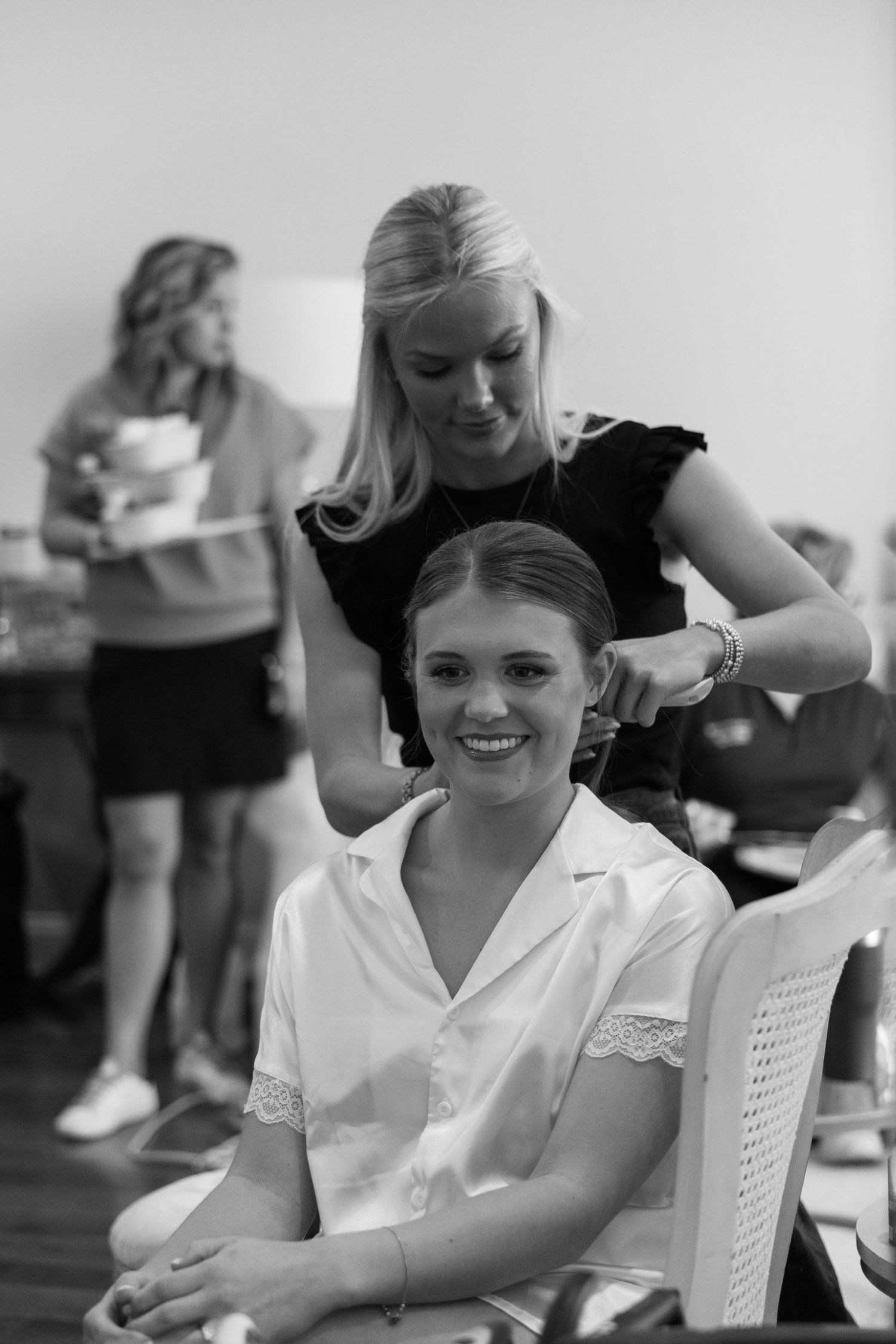 Bride smiling in chair getting hair styled by blonde hairdresser in black dress during bridal preparation.