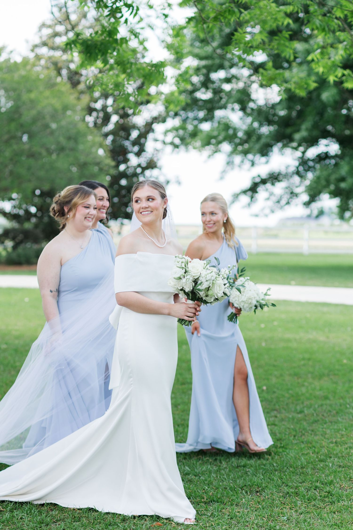 Bride in white off-shoulder gown holds bouquet while bridesmaids in blue dresses walk outdoors.