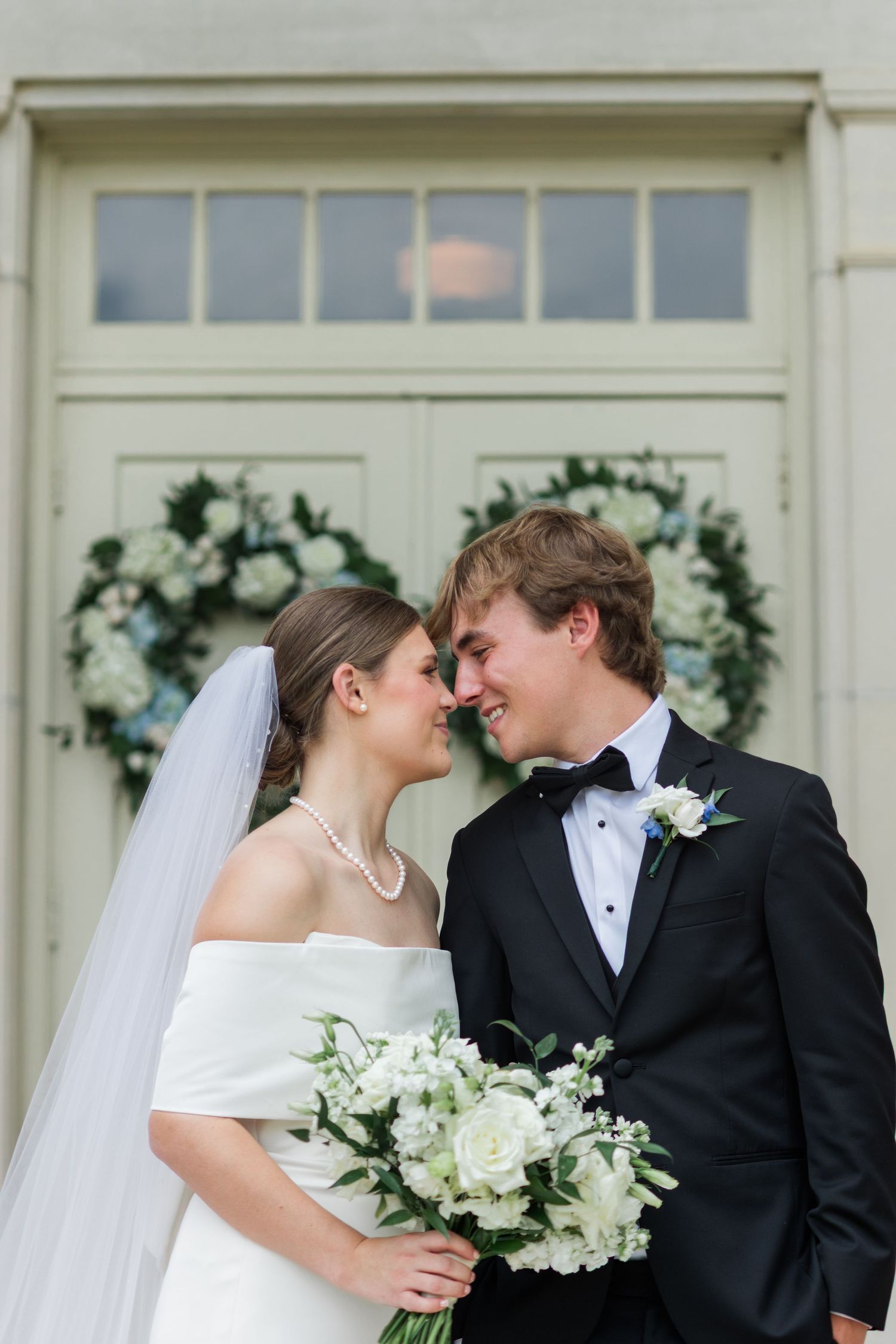 Bride and groom share intimate nose-to-nose moment before elegant doors with white floral wreaths.
