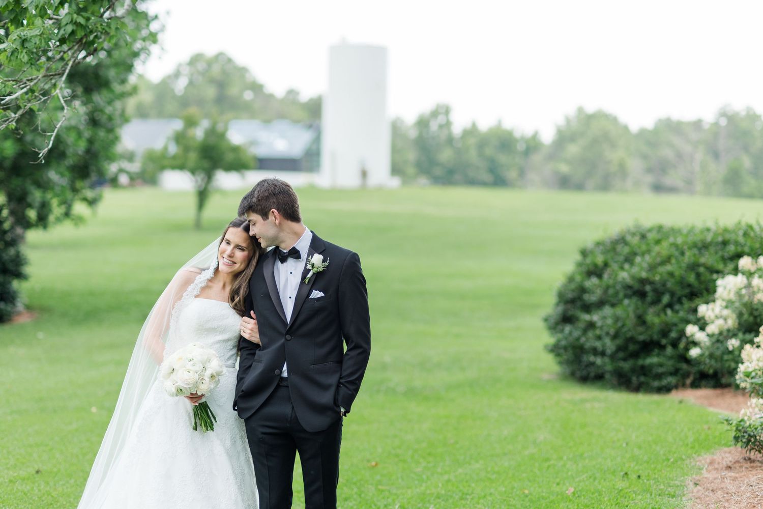 A bride and groom share a romantic moment on a lush green lawn with a white barn in the background.