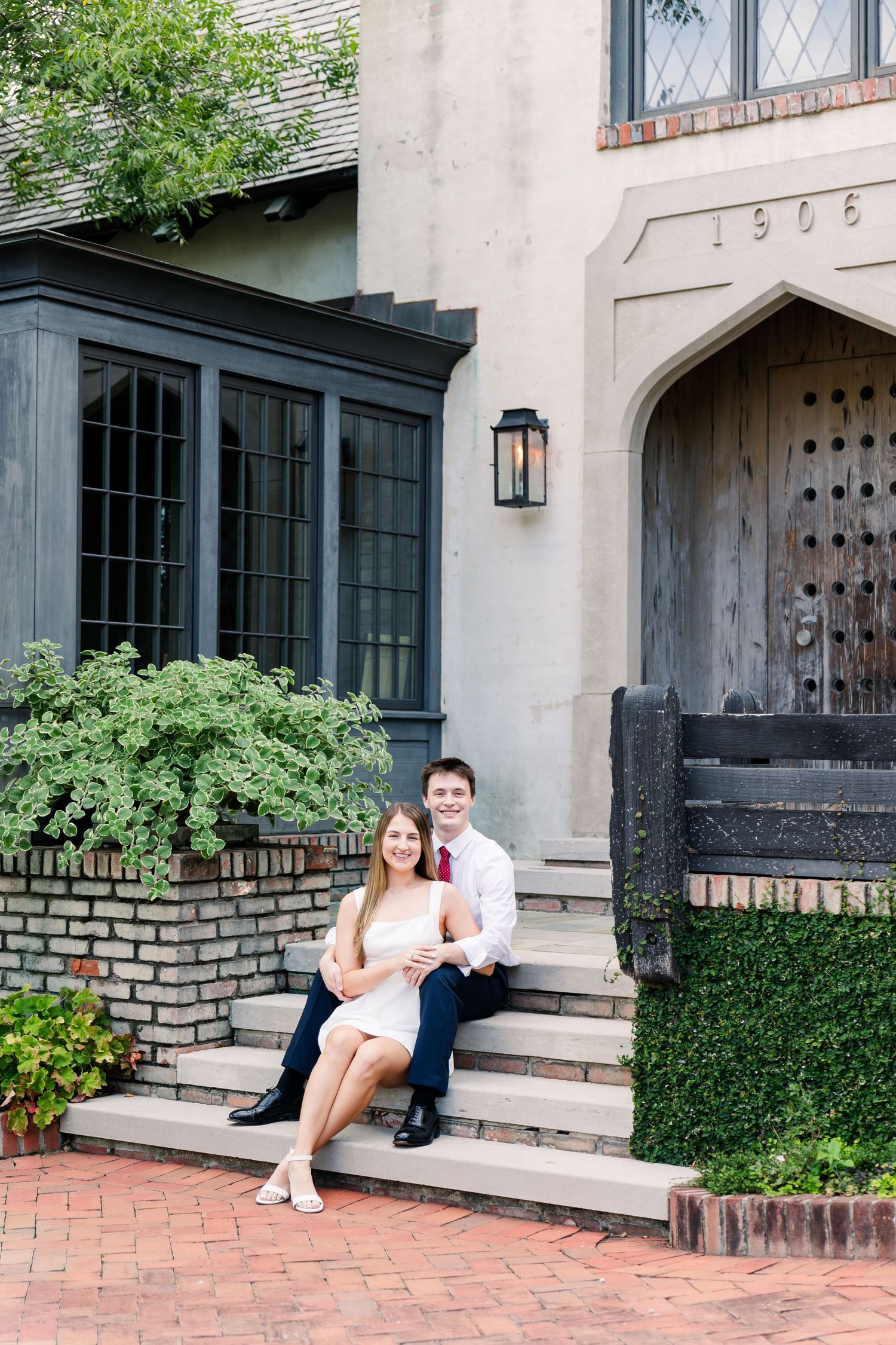 Couple sitting on brick steps outside a historic 1906 building, woman in white dress, man in white shirt and red tie.