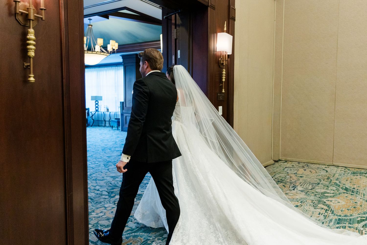 Bride and groom walking through elegant hotel hallway, her long lace wedding dress and cathedral veil trailing gracefully behind.