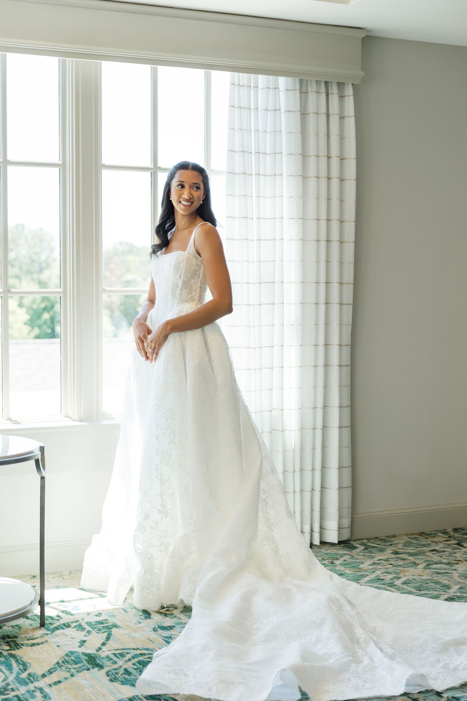 Smiling bride in a white lace A-line wedding gown with a long train standing by a bright window indoors.