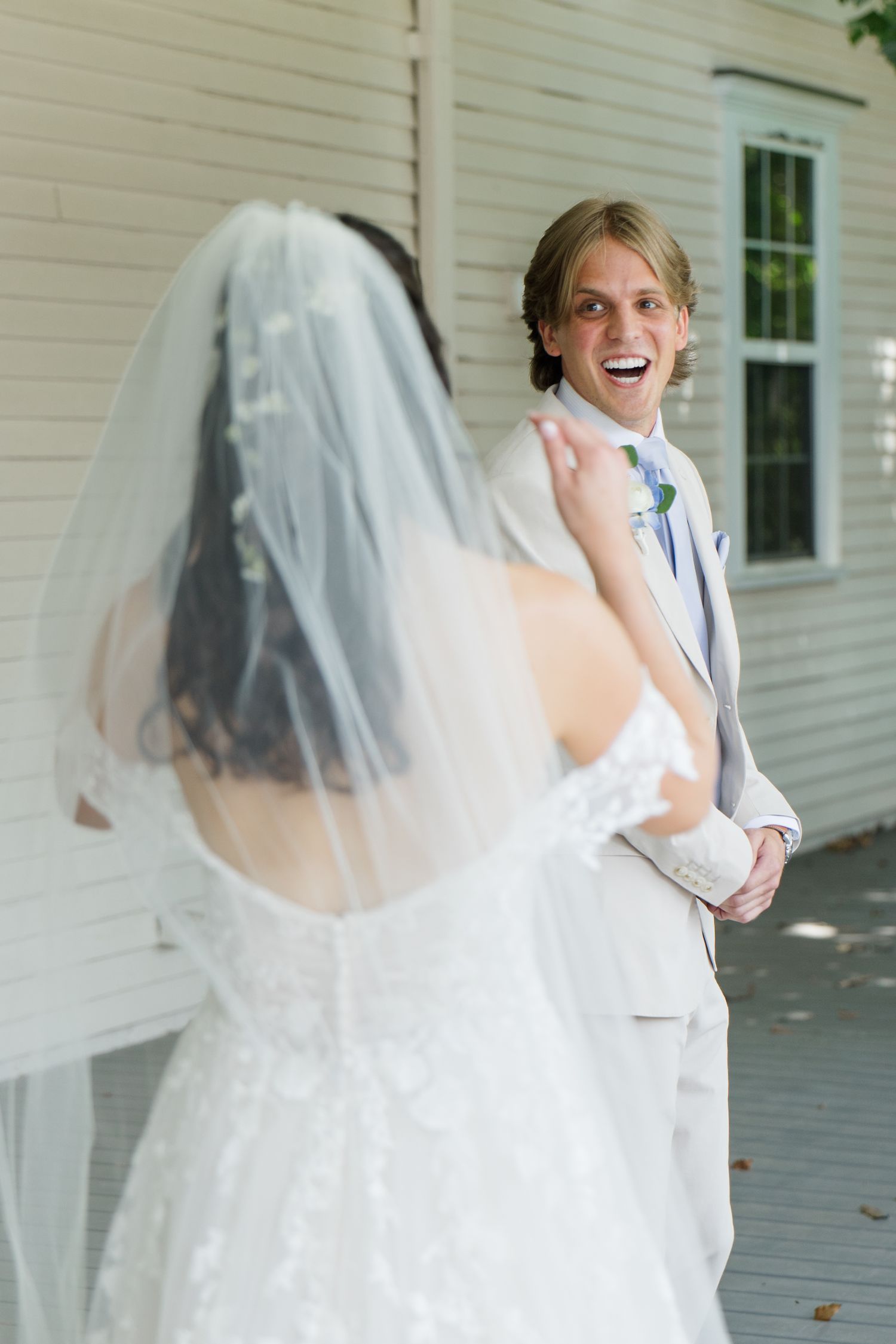 Groom in a tan suit laughing with joy during an emotional first look moment with his bride outdoors.
