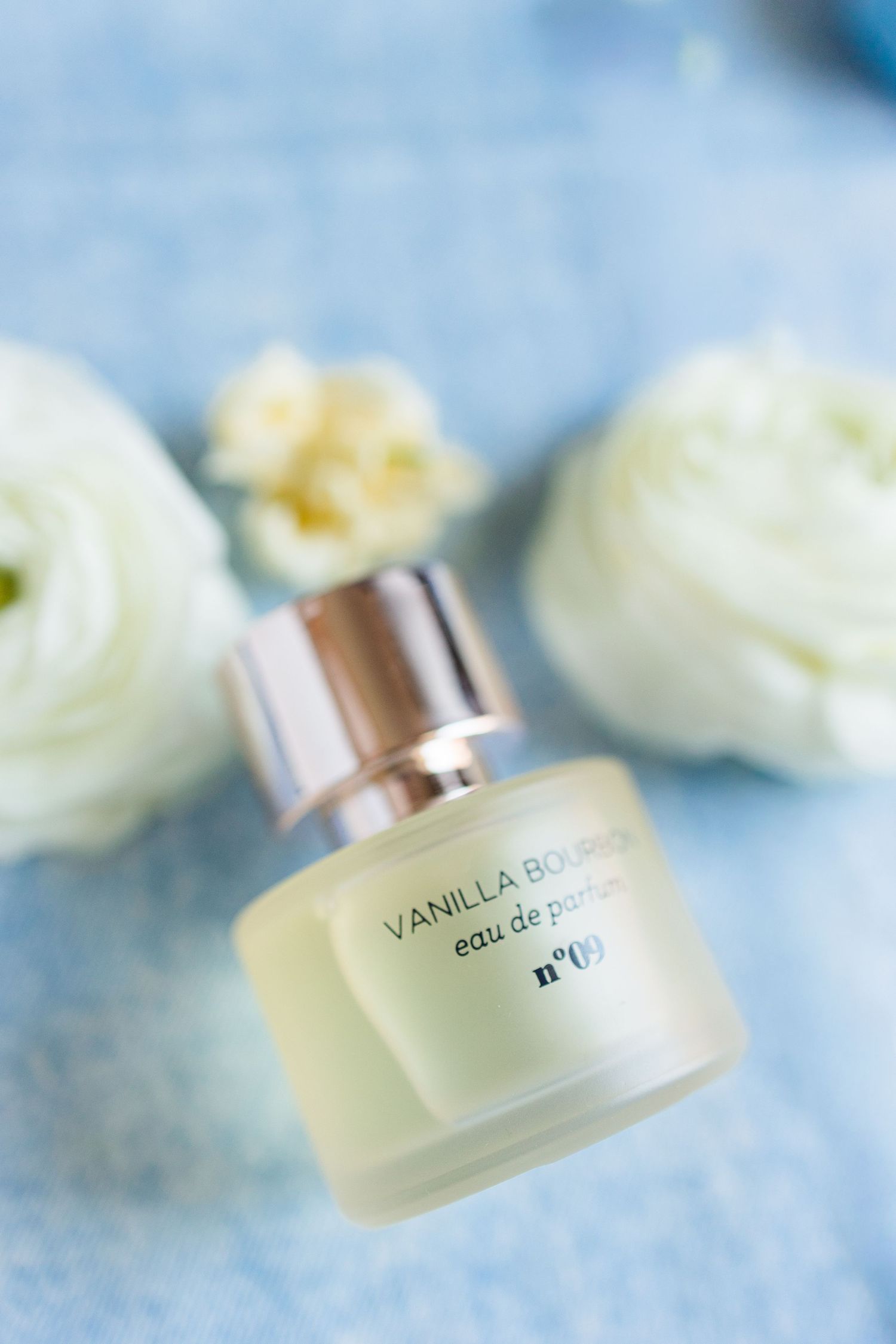 Small bottle of Vanilla Bouquet eau de parfum no.19 surrounded by white ranunculus flowers on blue background.