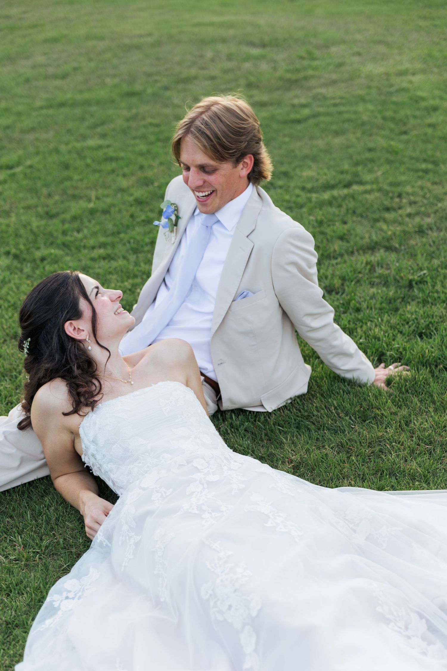 Laughing bride in white gown lying on grass with smiling groom in tan suit at wedding.