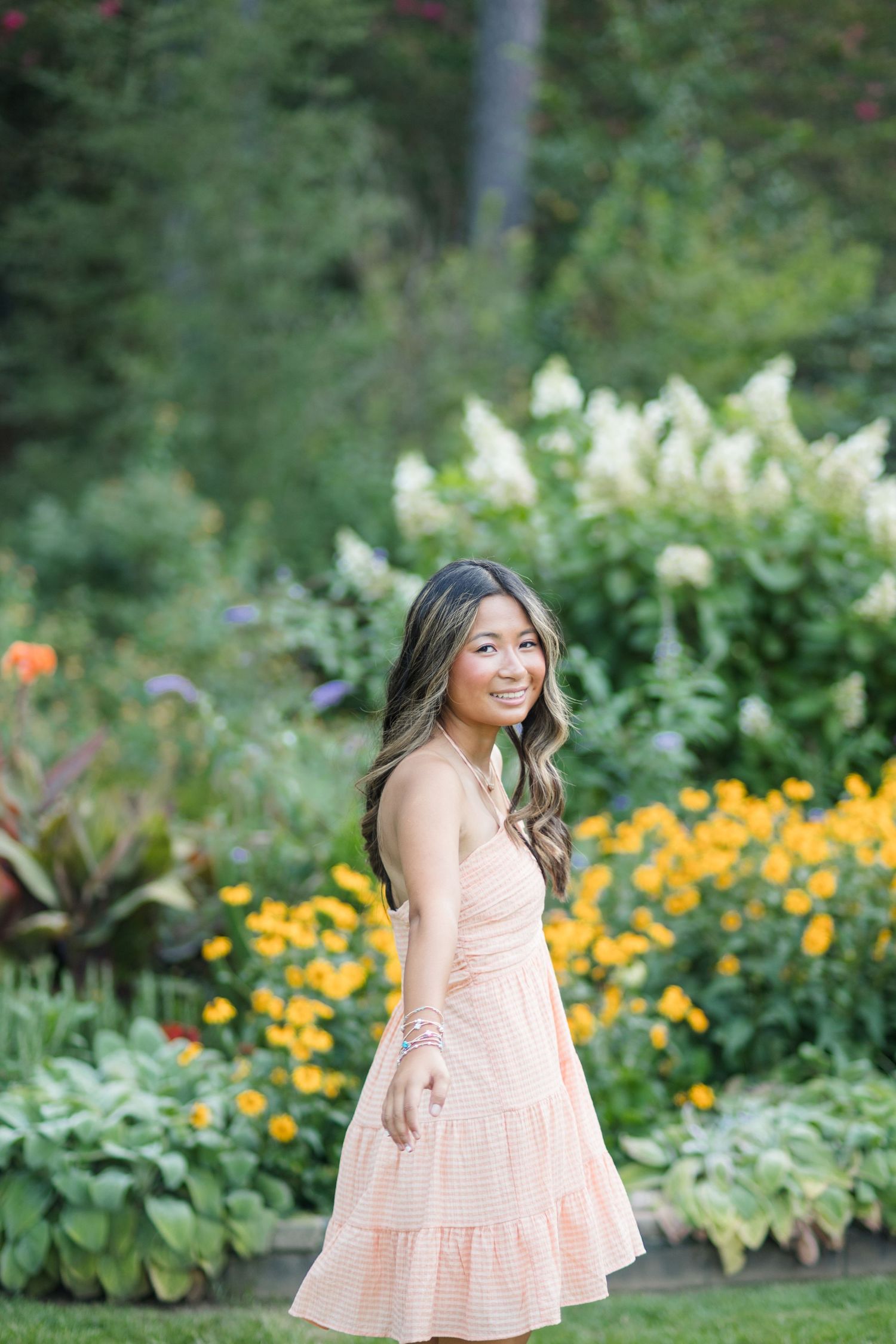 Smiling young woman in peach sundress posing in colorful garden with yellow flowers.