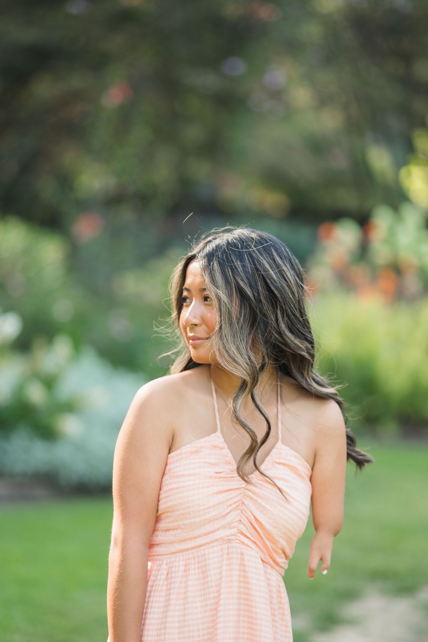 Young woman in a peach striped sundress poses outdoors in a lush green garden, her long wavy hair flowing.