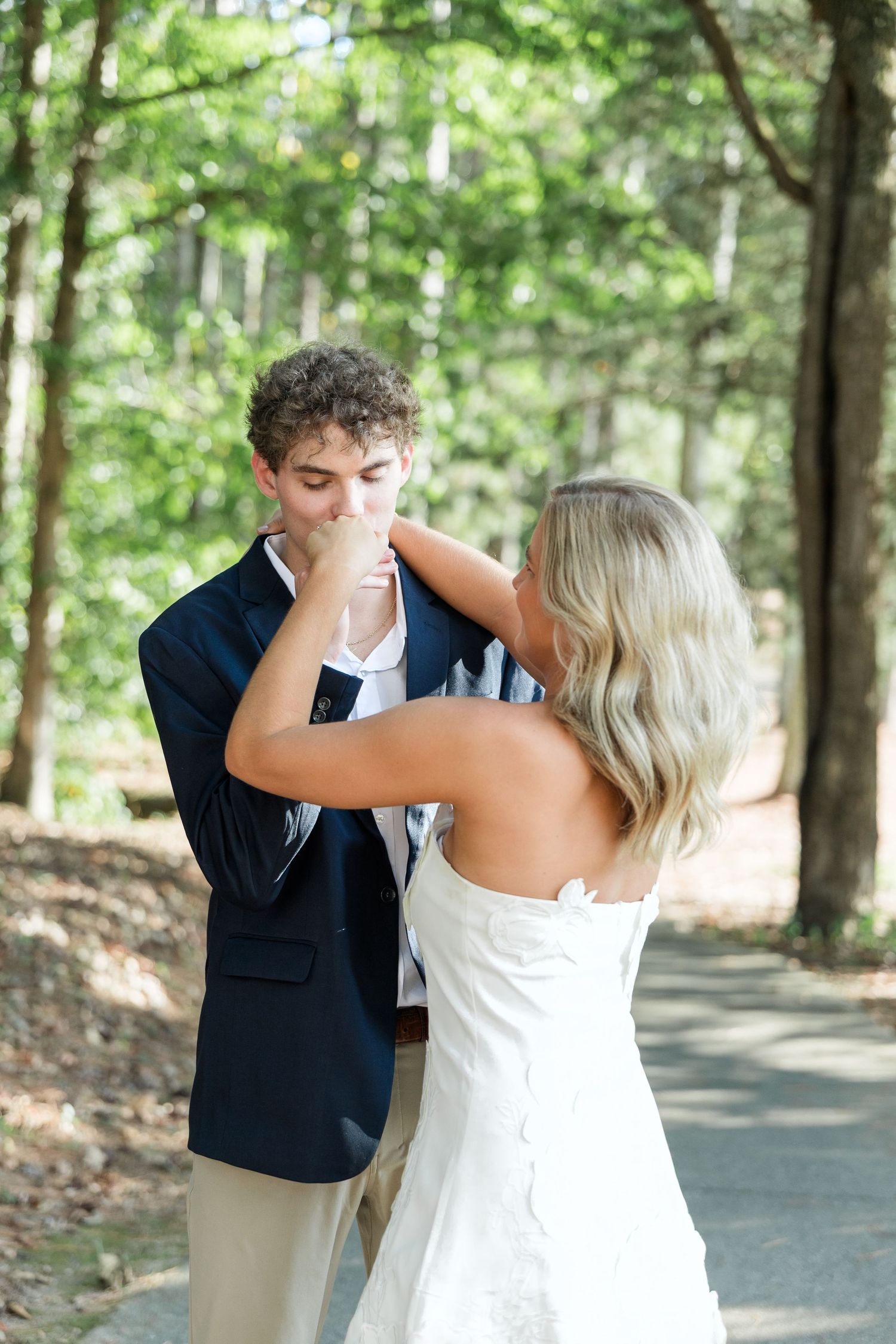 Groom kisses bride's hand tenderly on a sunlit forest path during wedding portraits.