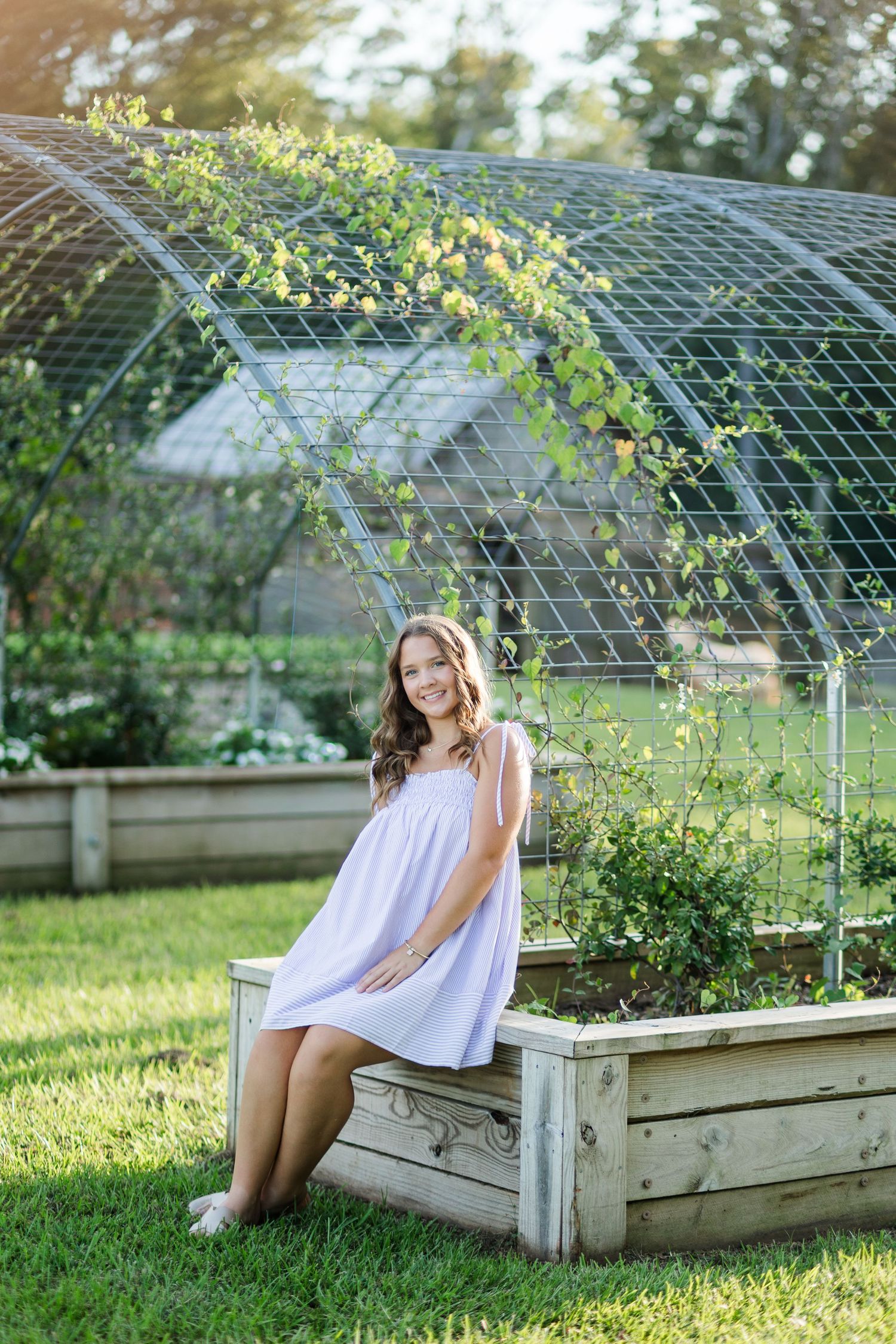 Young woman in white dress sitting on a raised garden bed beside an arched trellis outdoors.