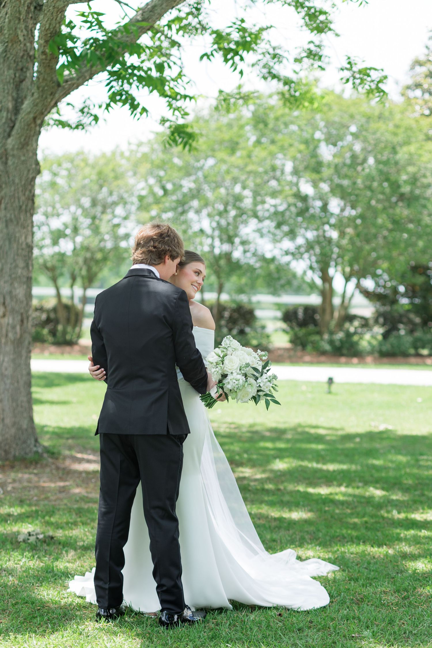Bride in white off-shoulder gown holds bouquet while embracing groom in black tuxedo under a tree outdoors.