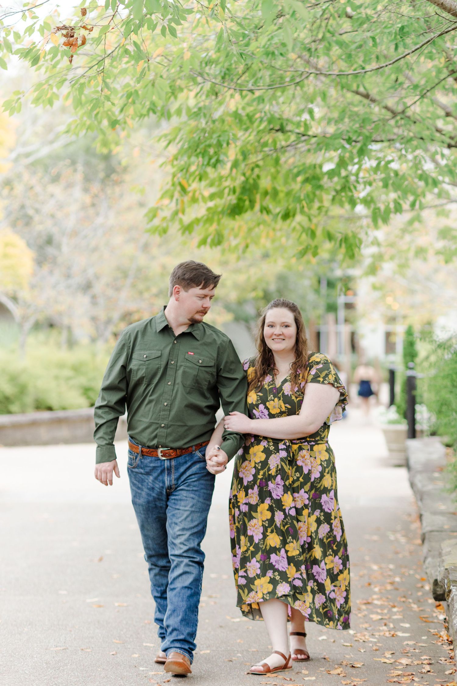 A couple walks hand-in-hand on a tree-lined path, smiling at each other, wearing a green shirt and floral dress.