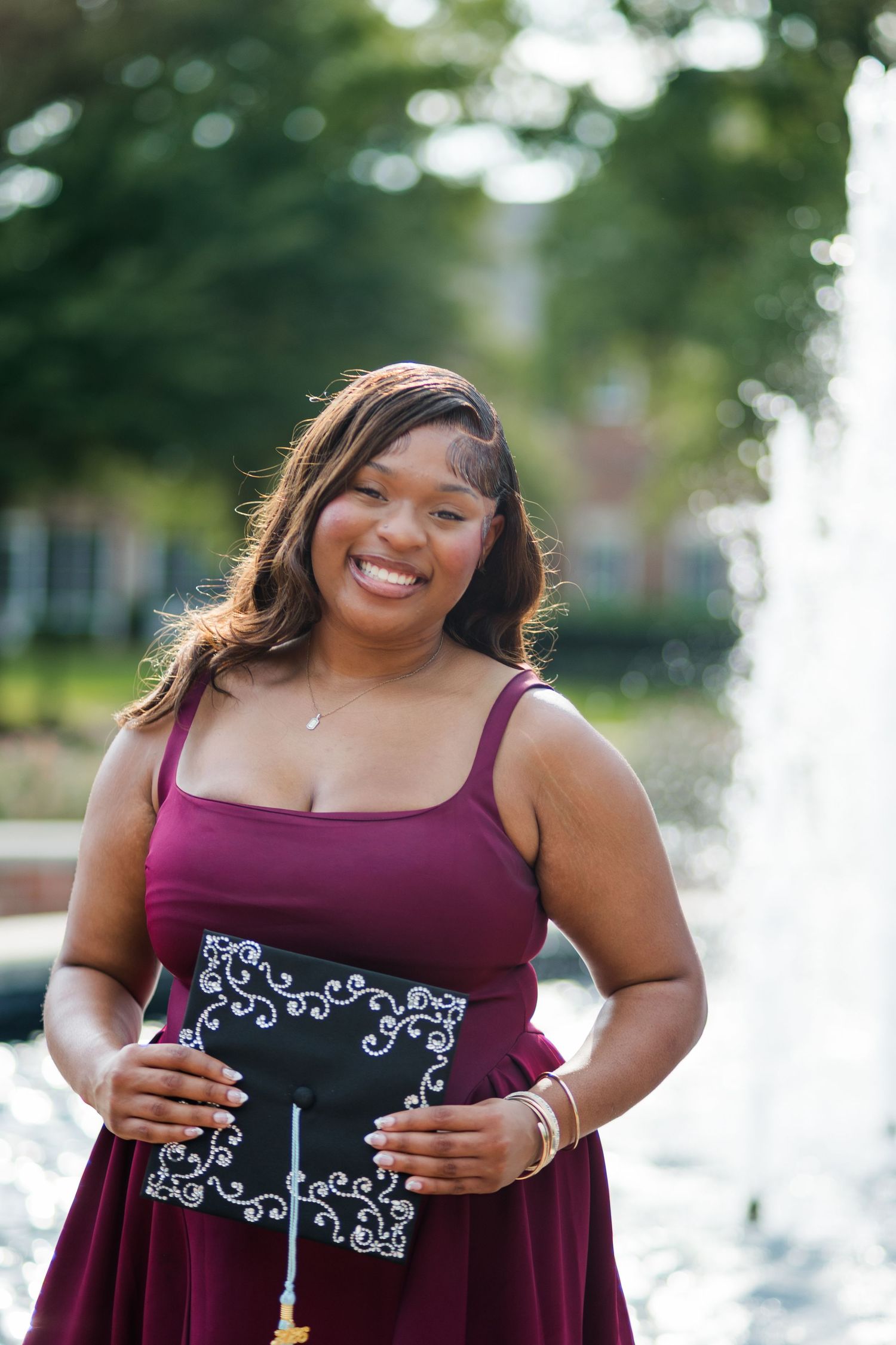 Smiling graduate in burgundy dress holding decorated mortarboard cap outdoors near fountain.