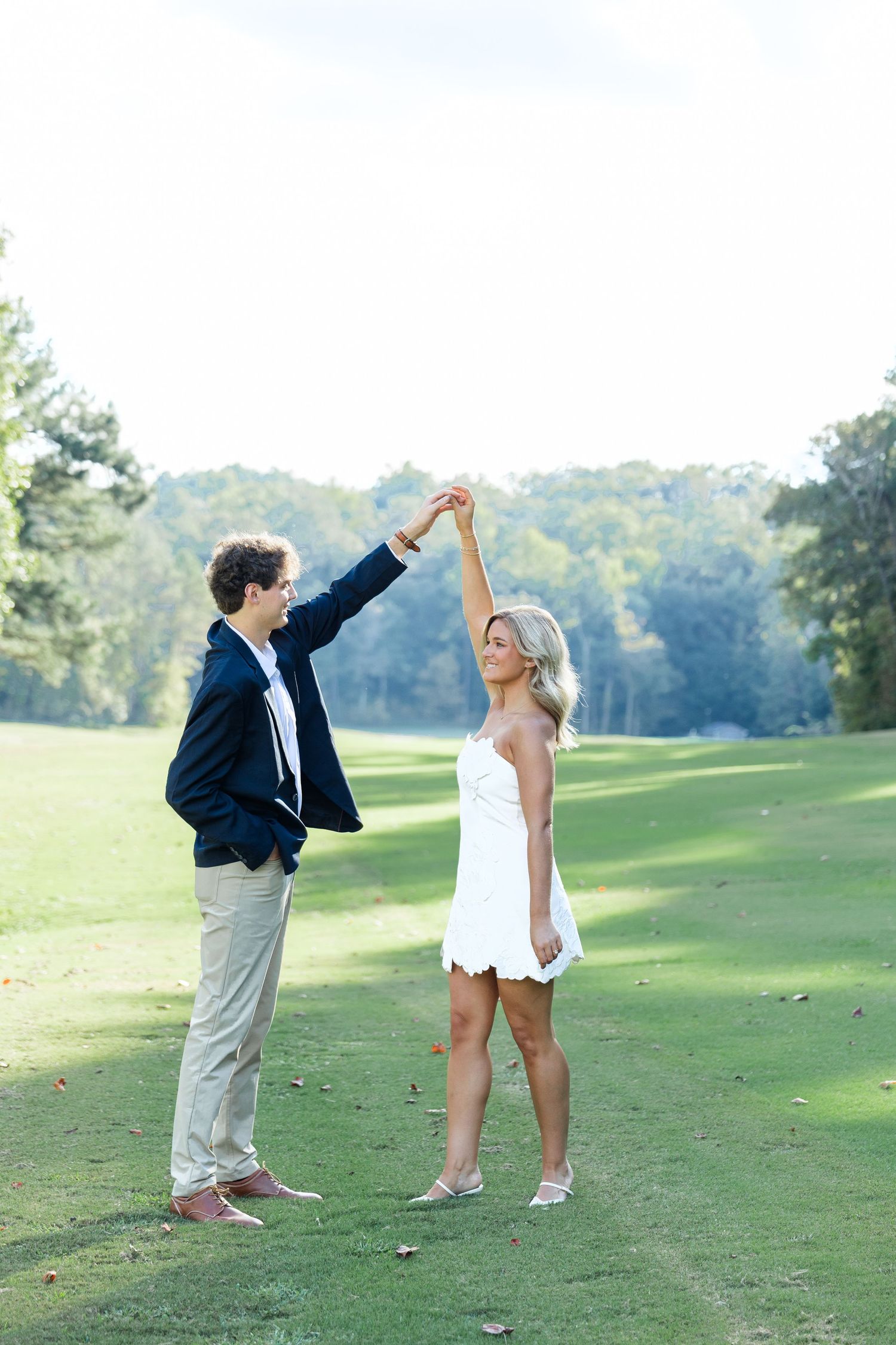 Couple dancing on golf course, man in navy blazer spinning woman in white mini dress outdoors.