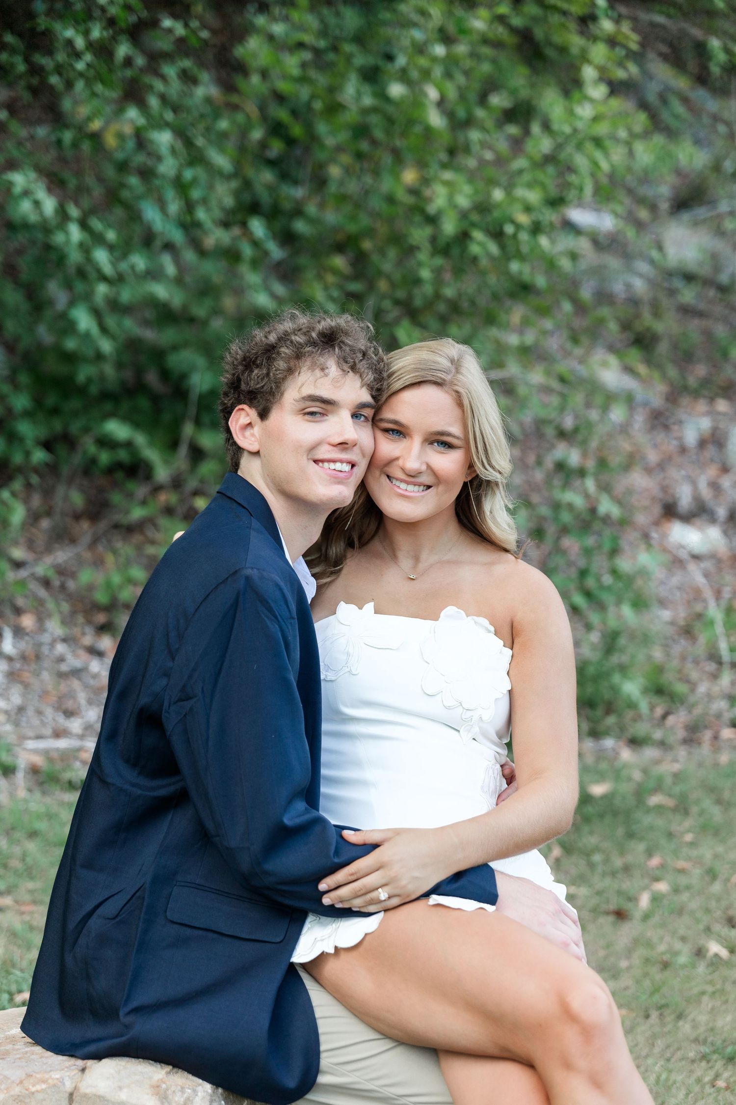 Smiling young couple sitting together outdoors on rocks with green foliage in the background.