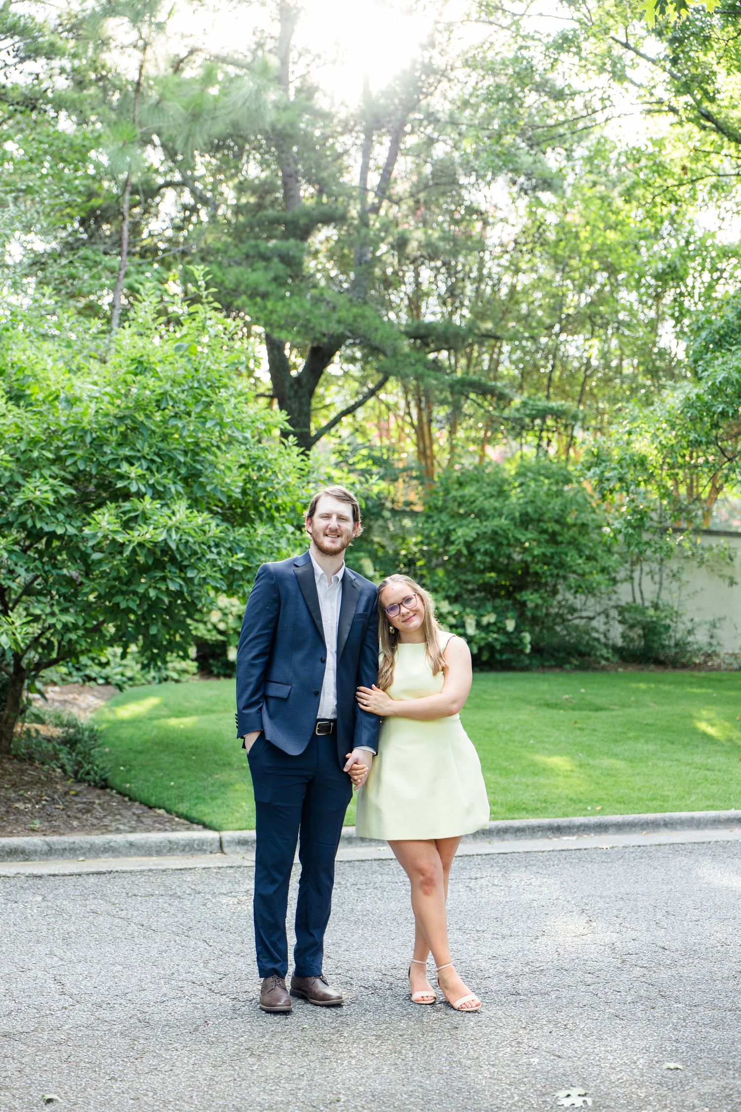 Smiling couple standing together outdoors, woman in mint dress and man in navy suit, surrounded by green trees.
