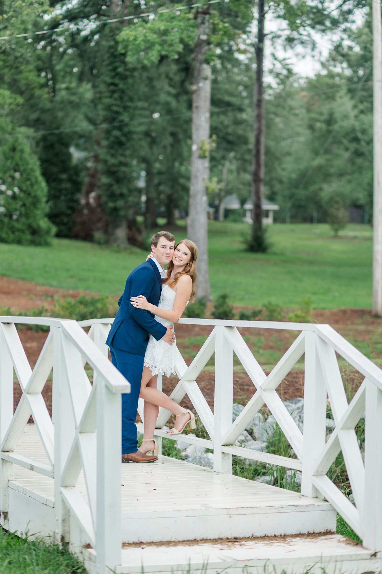 Happy couple posing together on a white decorative bridge surrounded by green trees and gardens.