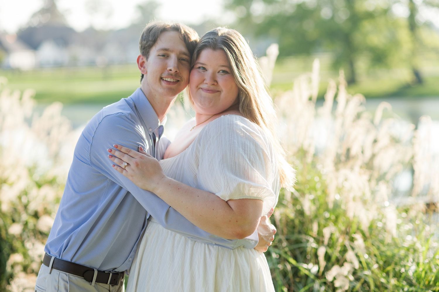 A couple embraces and smiles outdoors near tall grasses and a pond in warm golden sunlight.