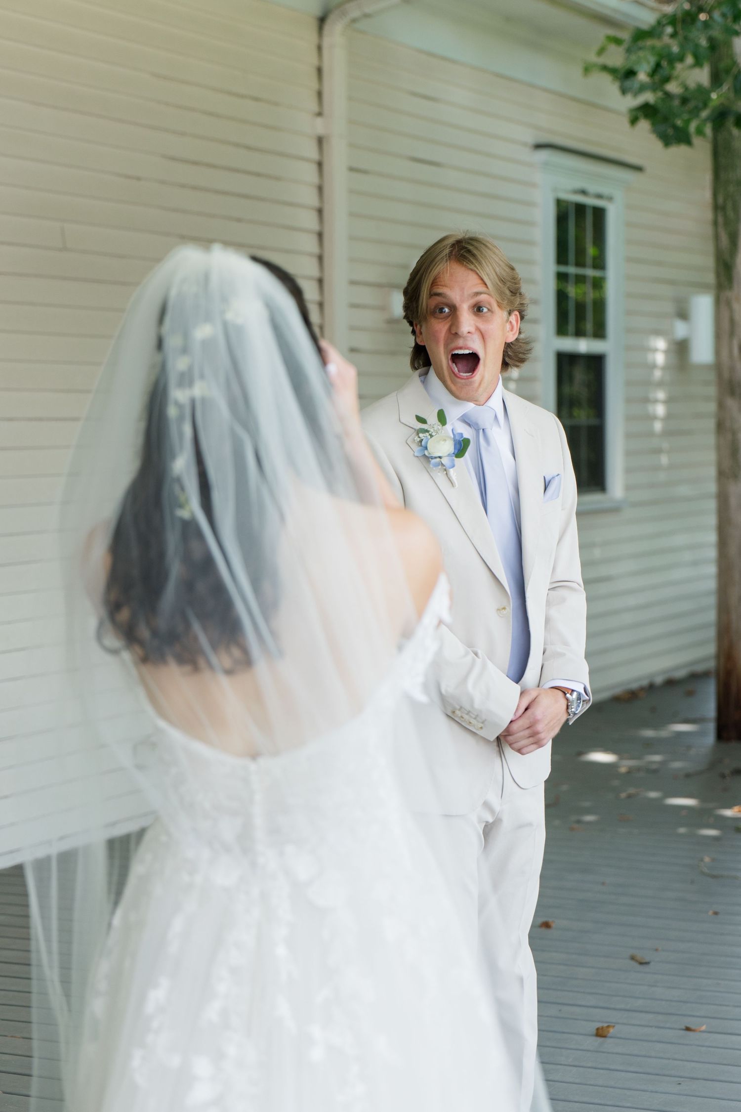 Groom in beige suit reacts with joyful surprise seeing bride in white lace gown and veil during first look.