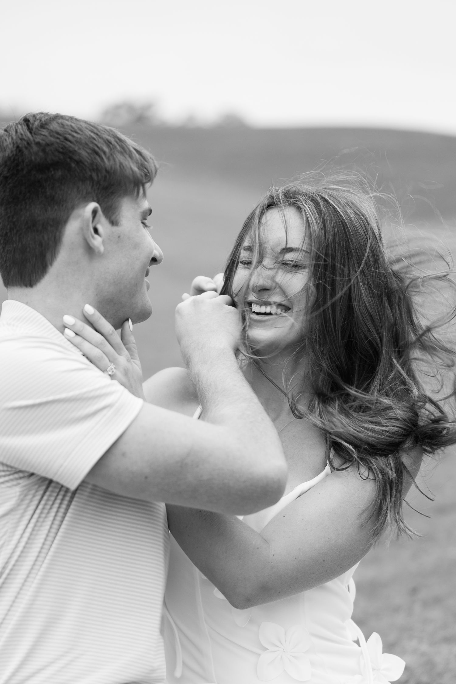 Black and white photo of a laughing woman with windblown hair playfully touching her partner's face during a joyful outdoor couple session.