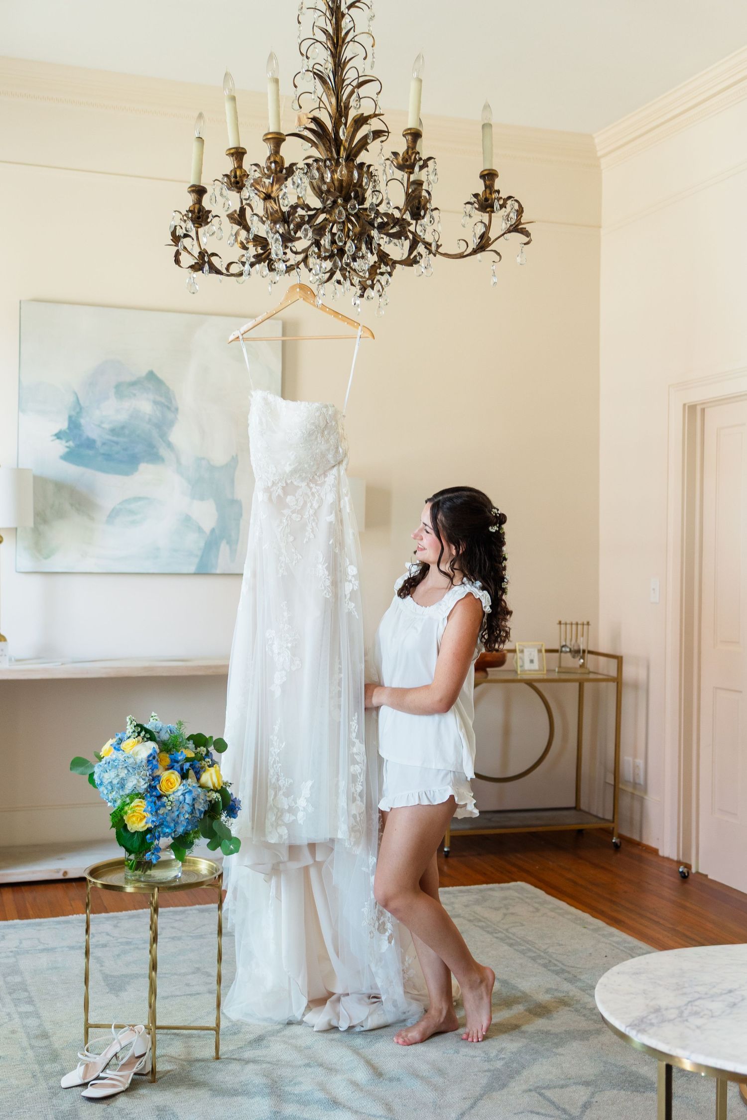 Bride in white robe admiring her lace wedding gown hanging from a chandelier, with a blue and yellow floral bouquet nearby.