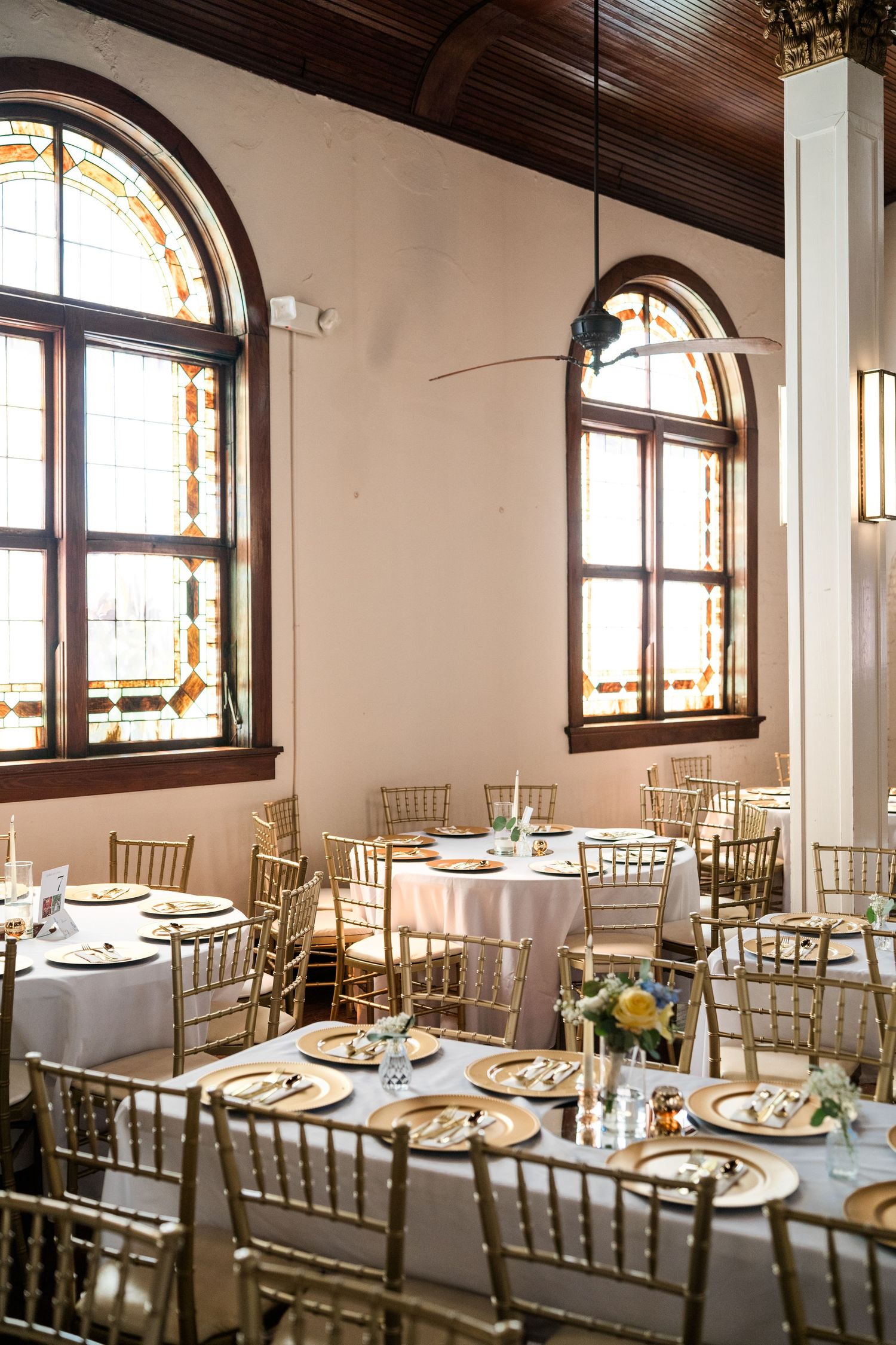 Elegant wedding reception hall with gold Chiavari chairs, white round tables, and arched stained glass windows.