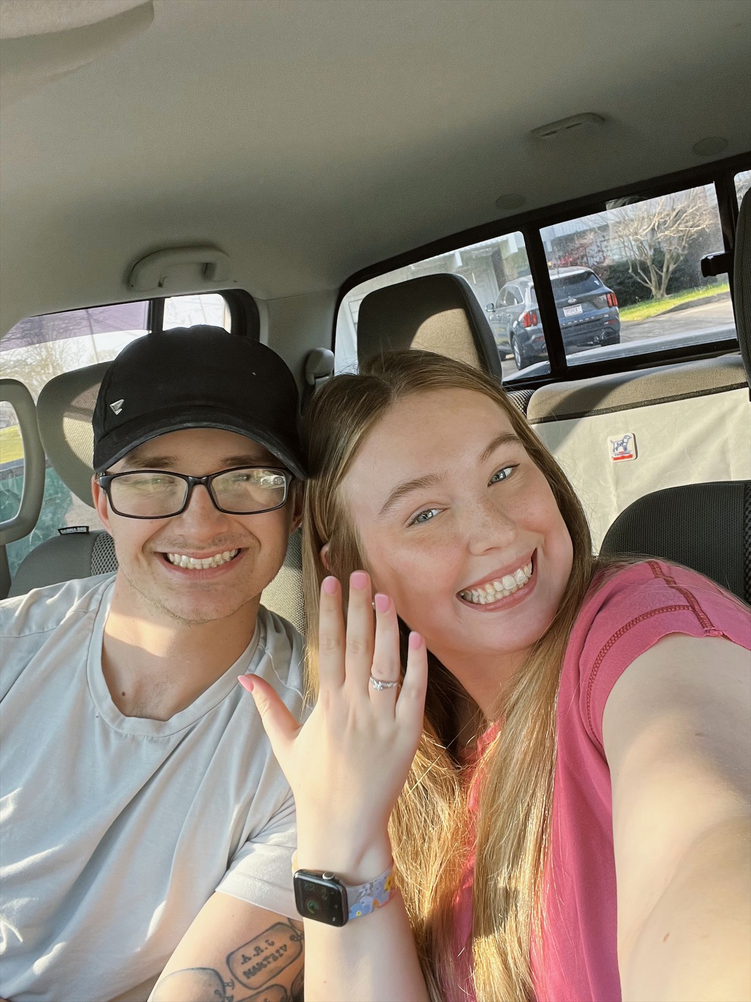 Smiling engaged couple in car showing off engagement ring in joyful selfie photo.