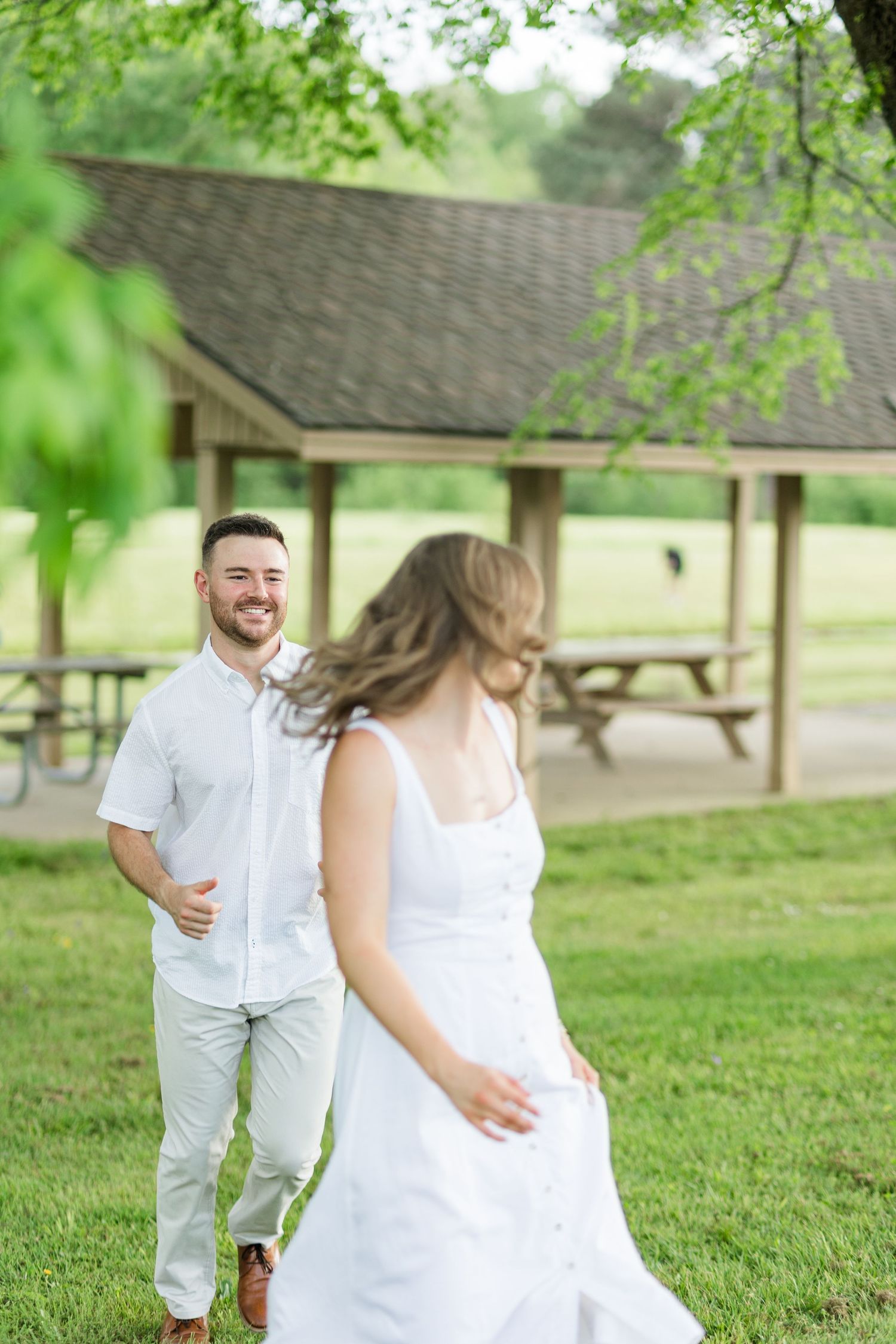 Smiling man in white shirt chasing laughing woman in white dress across green park lawn.
